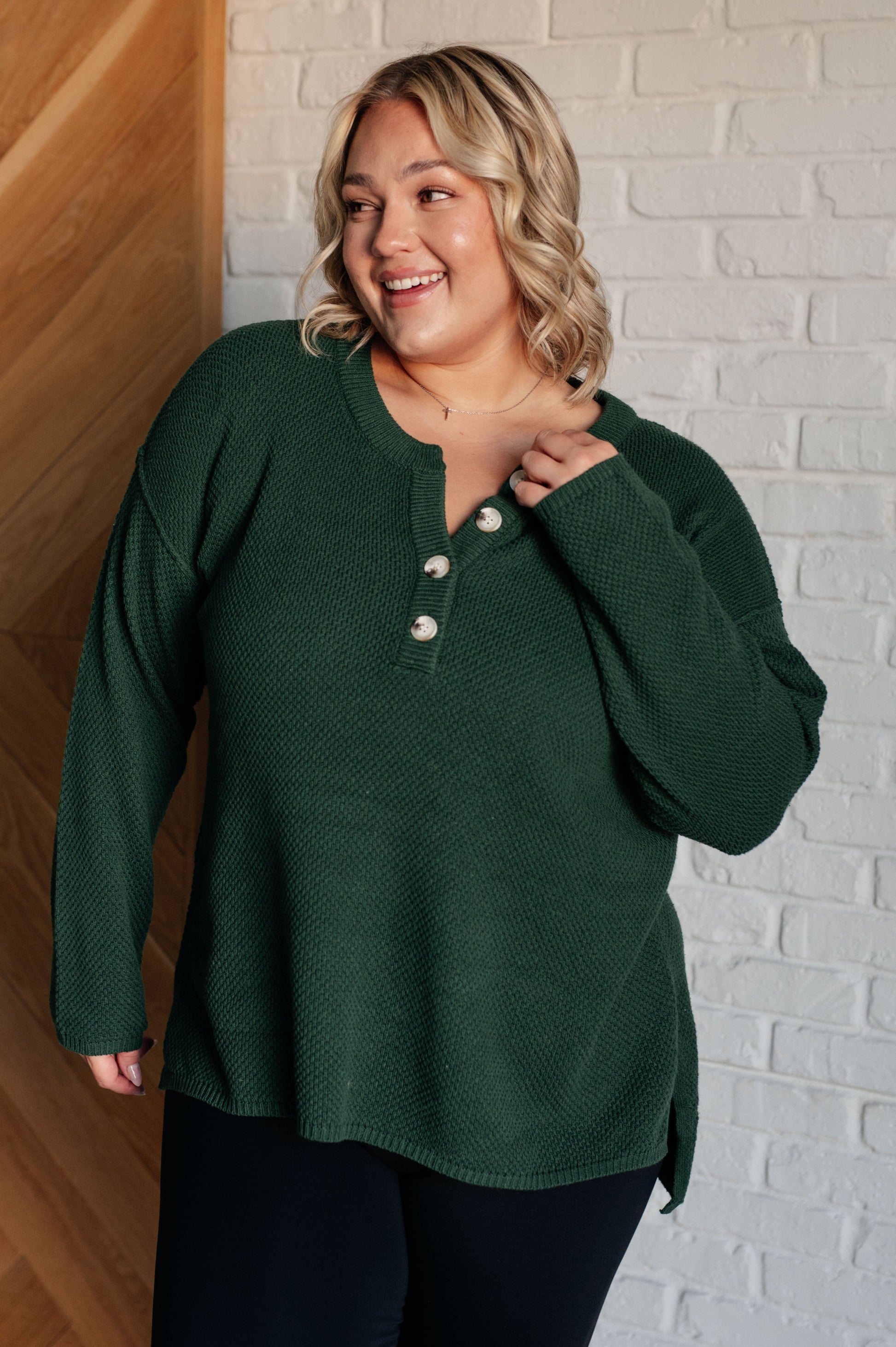 Women's dark green henley sweater with a ribbed button-up neckline, exposed seams, long sleeves, and a stepped hem with side slits.