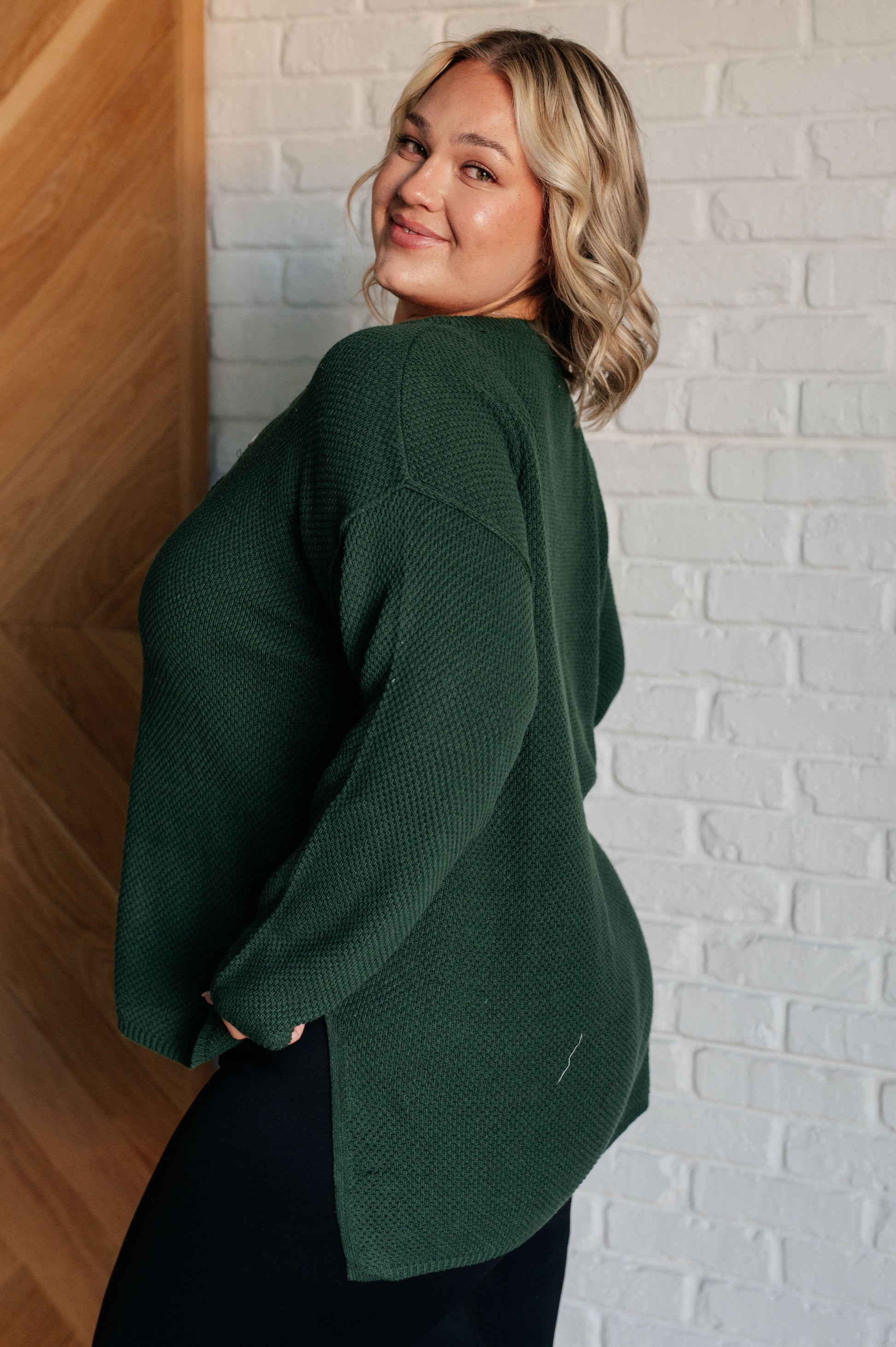 Women's dark green henley sweater with a ribbed button-up neckline, exposed seams, long sleeves, and a stepped hem with side slits.