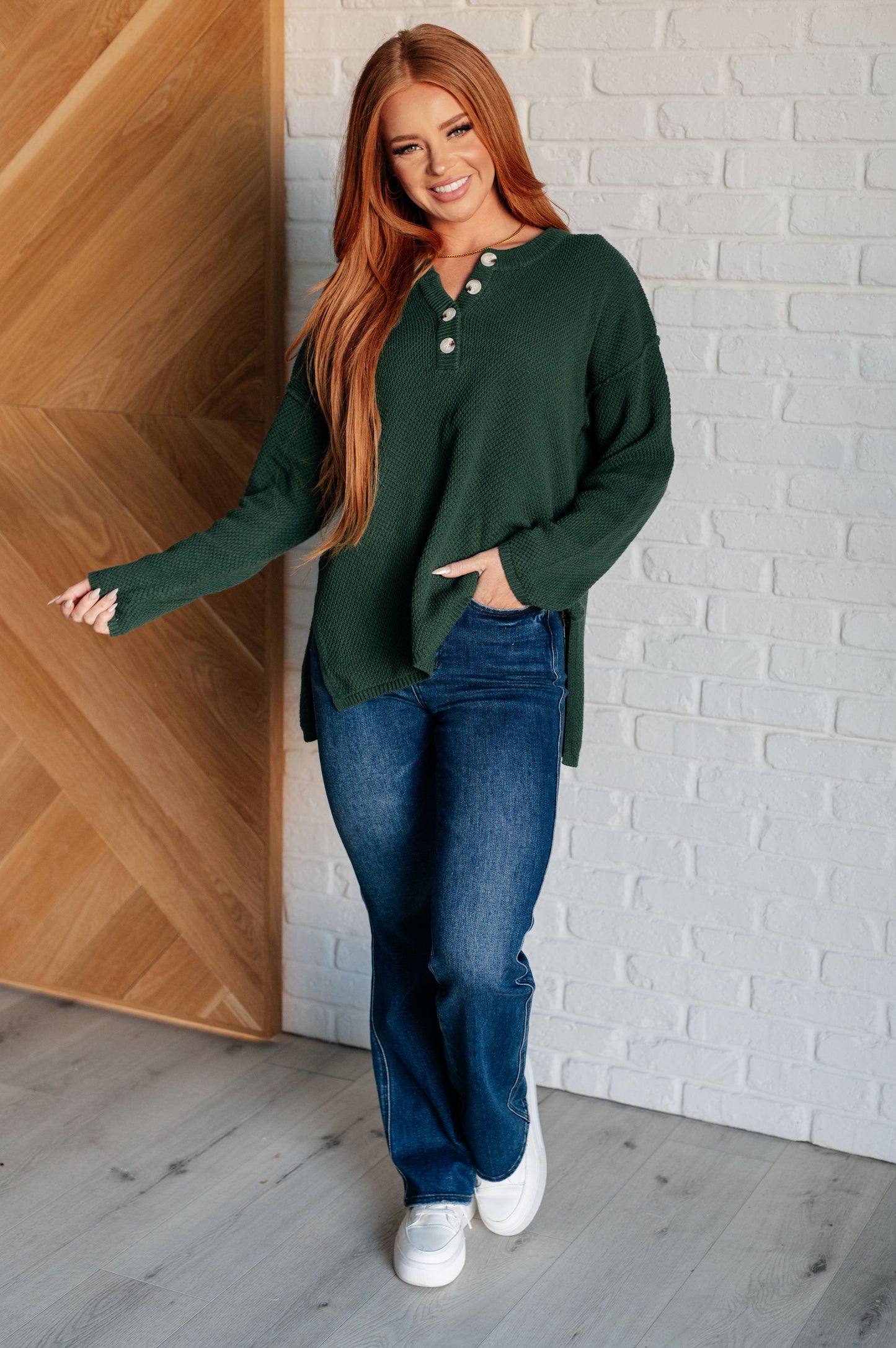 Women's dark green henley sweater with a ribbed button-up neckline, exposed seams, long sleeves, and a stepped hem with side slits.