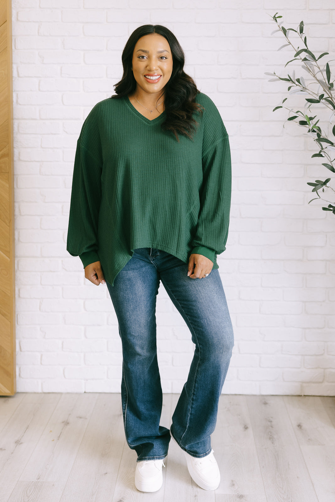 Women's green waffle knit top with a V-neckline, long sleeves, banded sleeve cuffs, faux tag detail, and a scooped asymmetrical hemline.