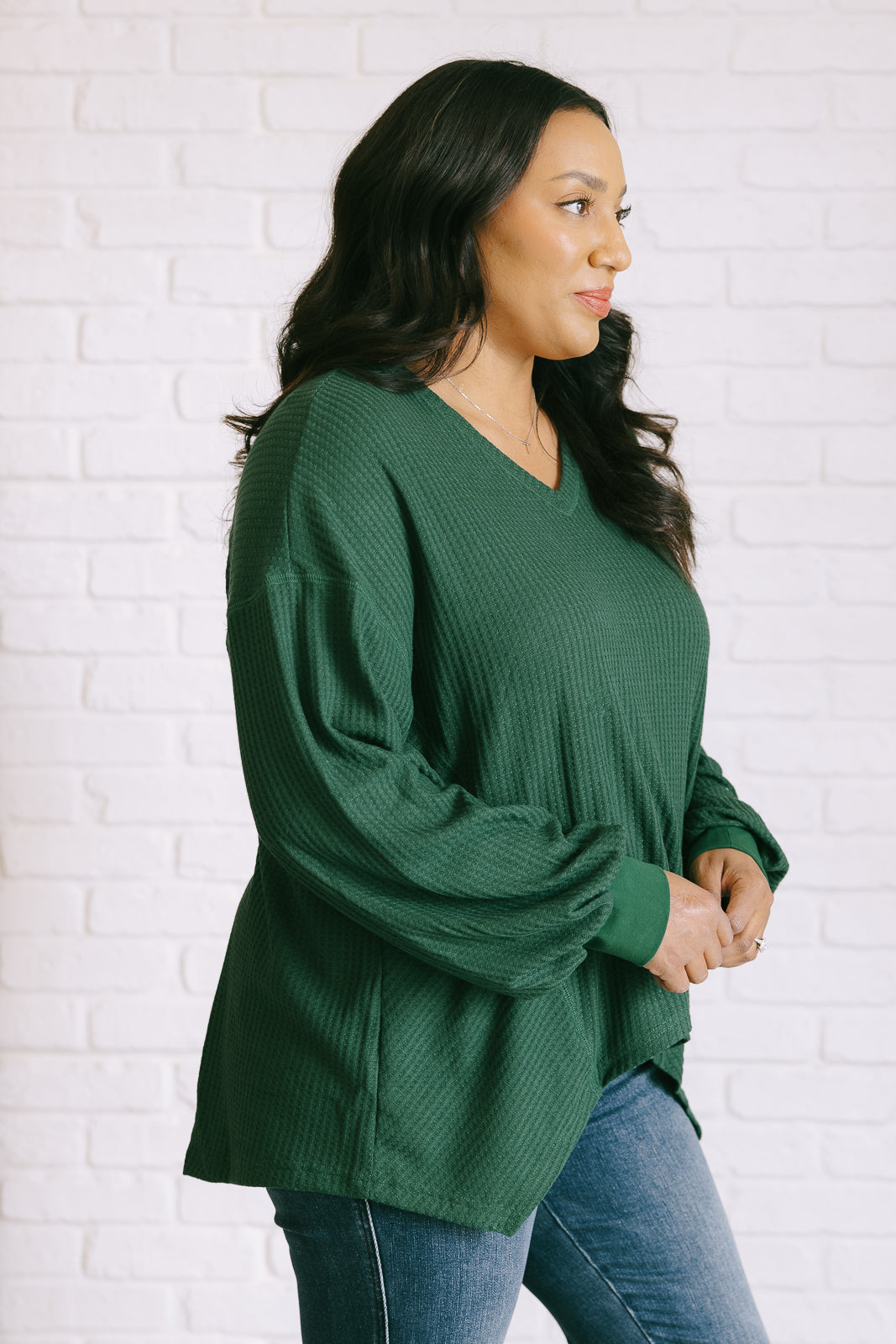 Women's green waffle knit top with a V-neckline, long sleeves, banded sleeve cuffs, faux tag detail, and a scooped asymmetrical hemline.