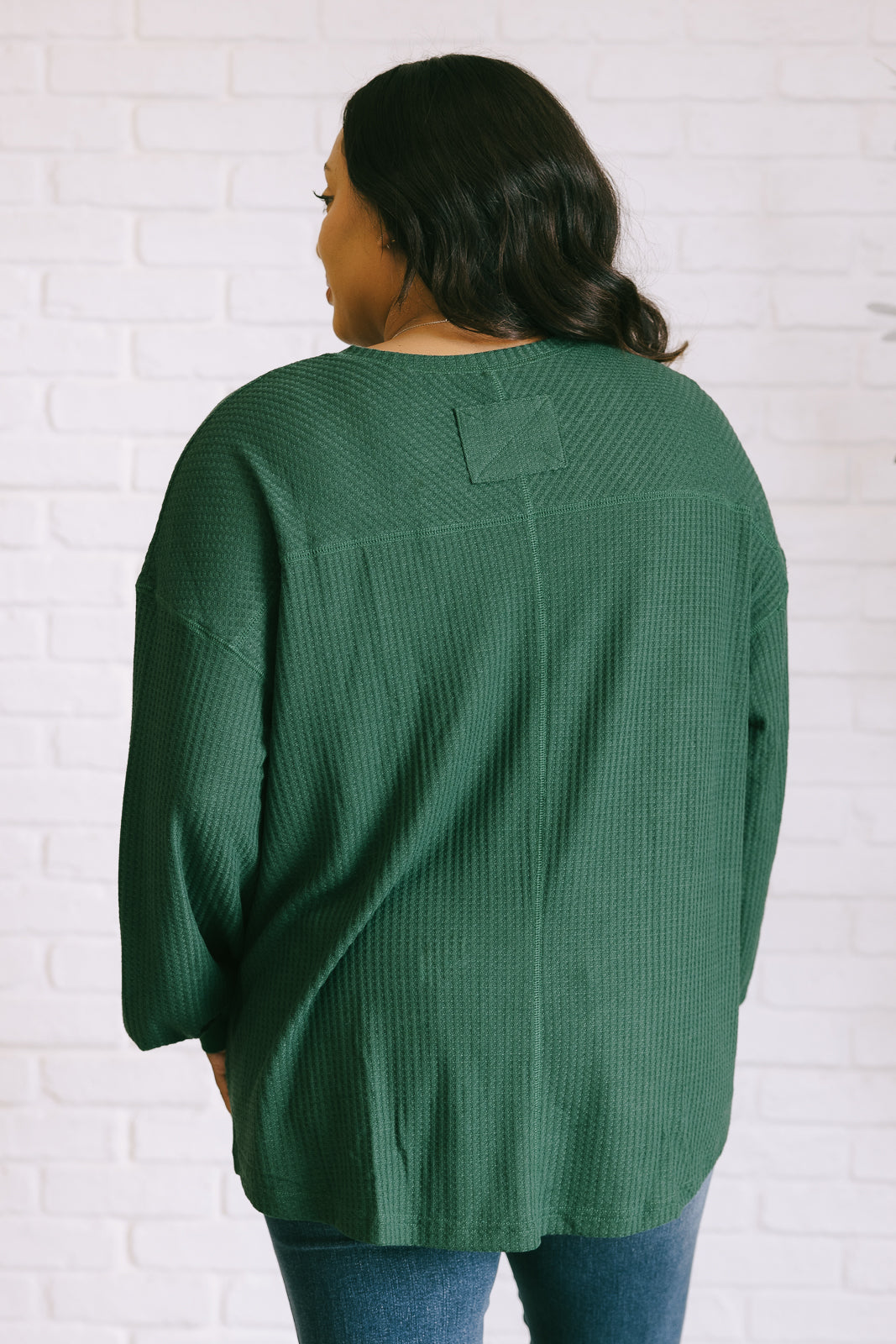 Women's green waffle knit top with a V-neckline, long sleeves, banded sleeve cuffs, faux tag detail, and a scooped asymmetrical hemline.