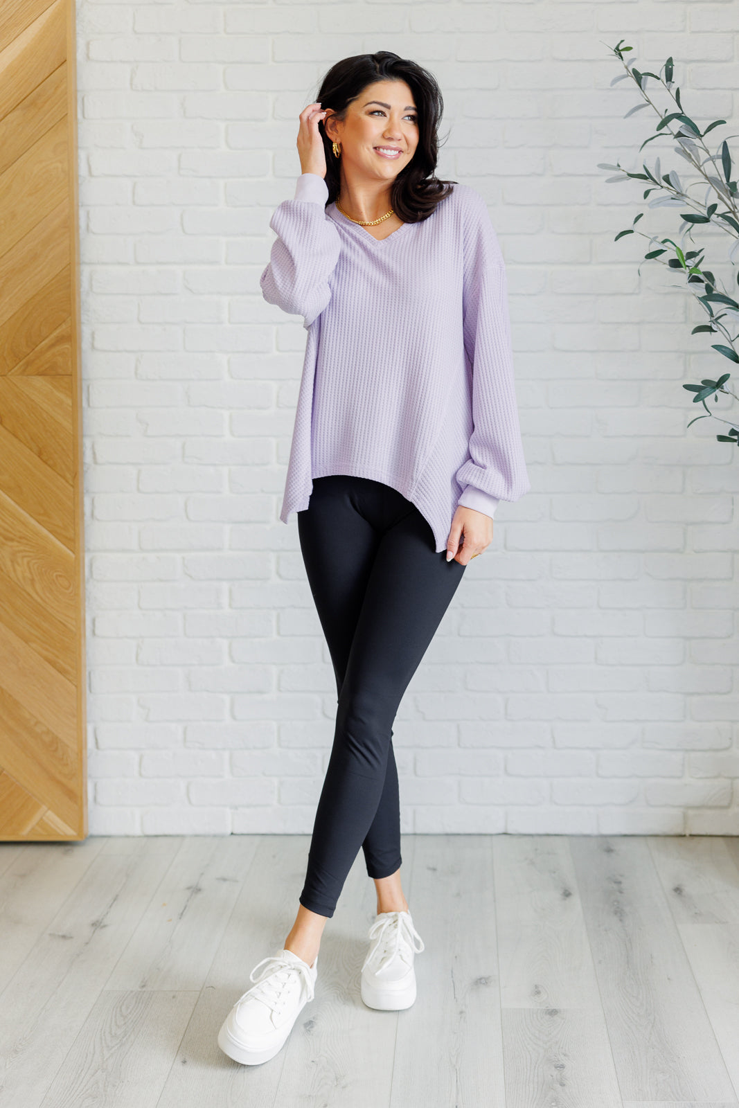 Women's lavender waffle knit top with a V-neckline, long sleeves, banded sleeve cuffs, faux tag detail, and a scooped asymmetrical hemline.