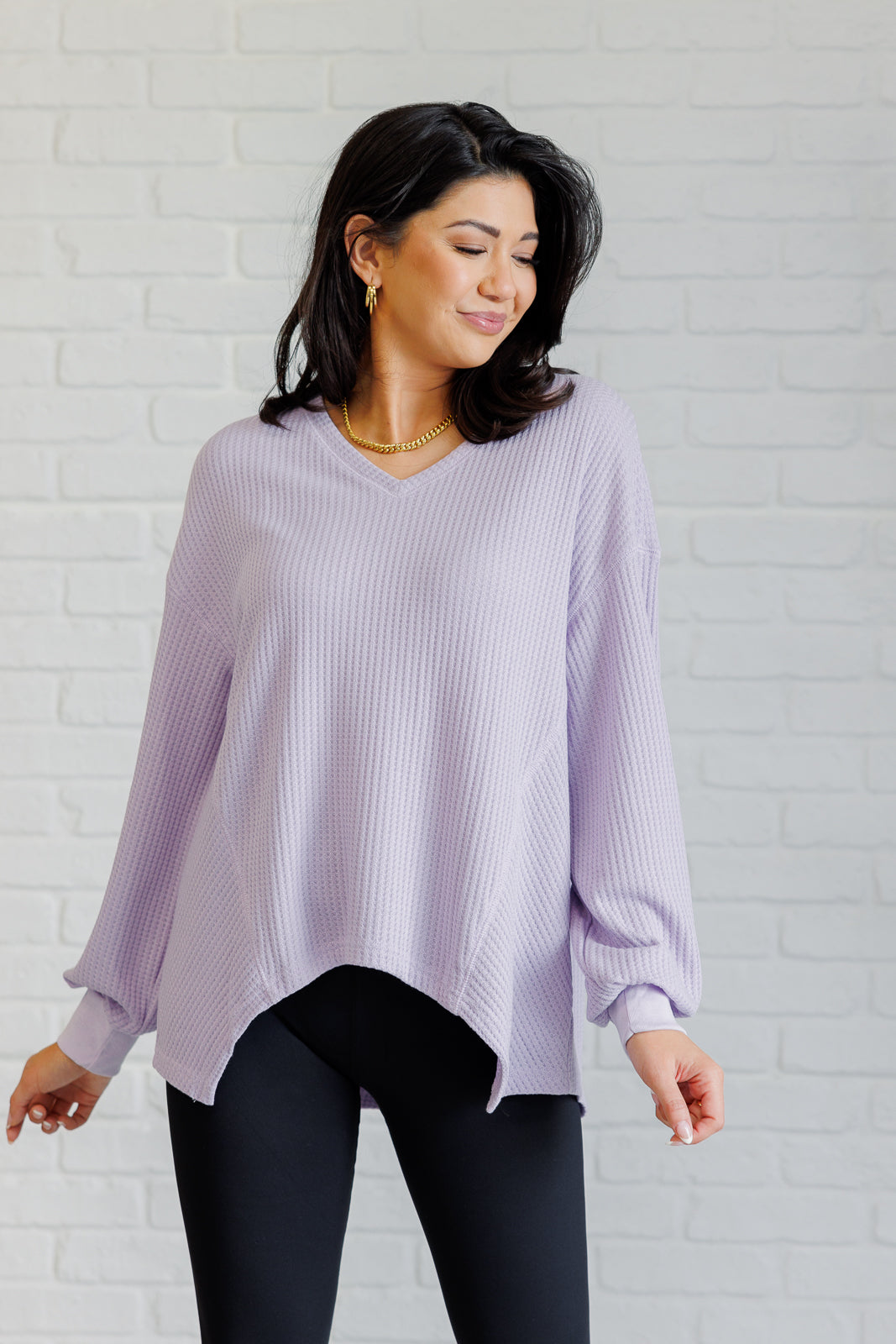 Women's lavender waffle knit top with a V-neckline, long sleeves, banded sleeve cuffs, faux tag detail, and a scooped asymmetrical hemline.