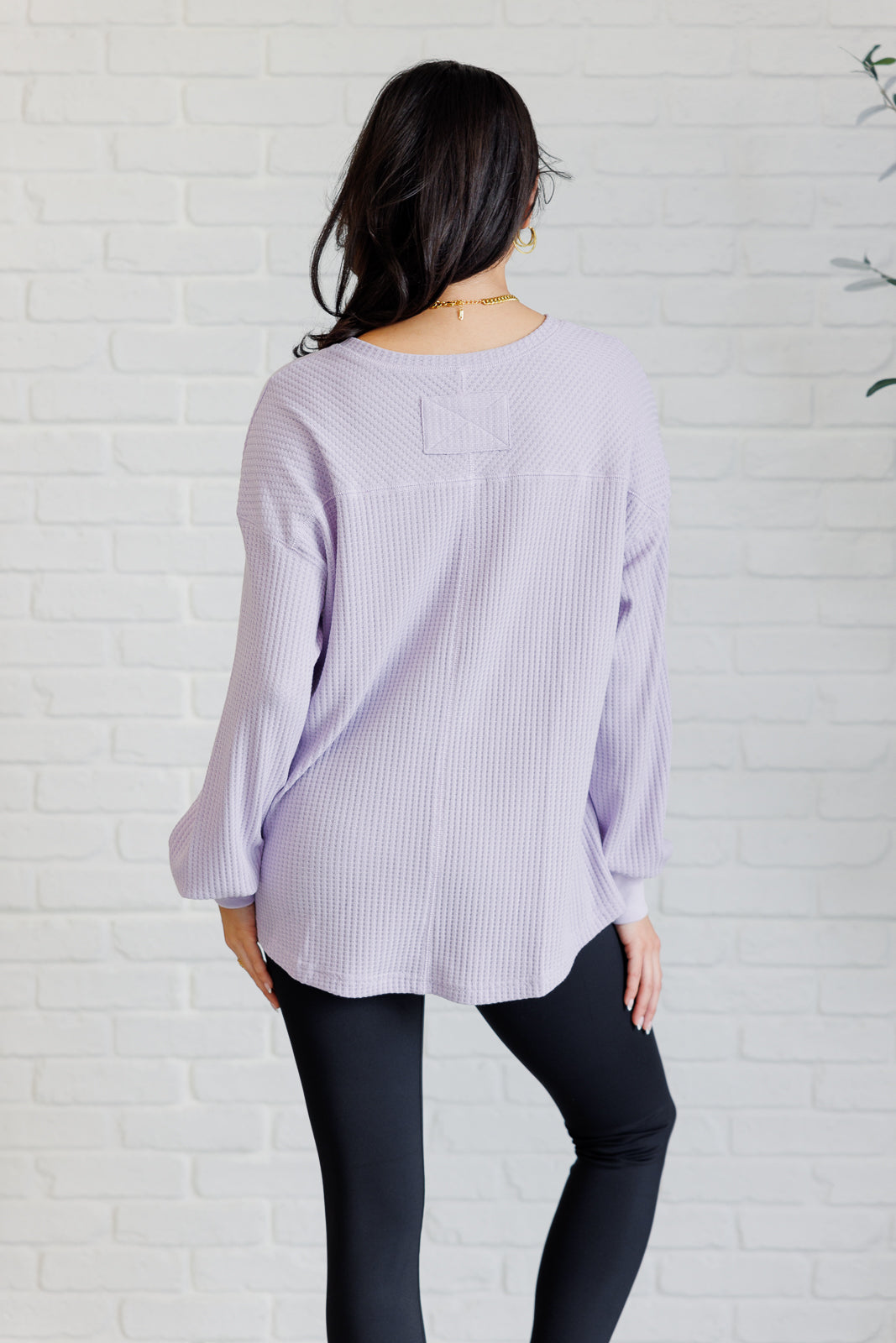 Women's lavender waffle knit top with a V-neckline, long sleeves, banded sleeve cuffs, faux tag detail, and a scooped asymmetrical hemline.