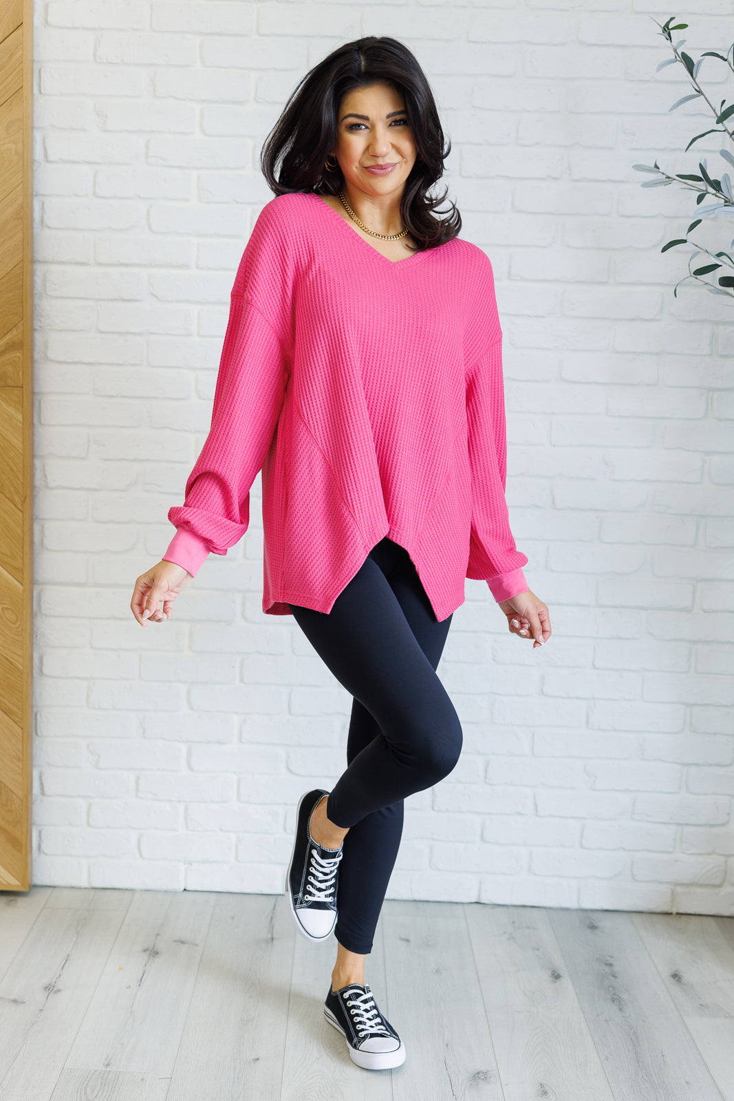 Women's pink waffle knit top with a V-neckline, long sleeves, banded sleeve cuffs, faux tag detail, and a scooped asymmetrical hemline.