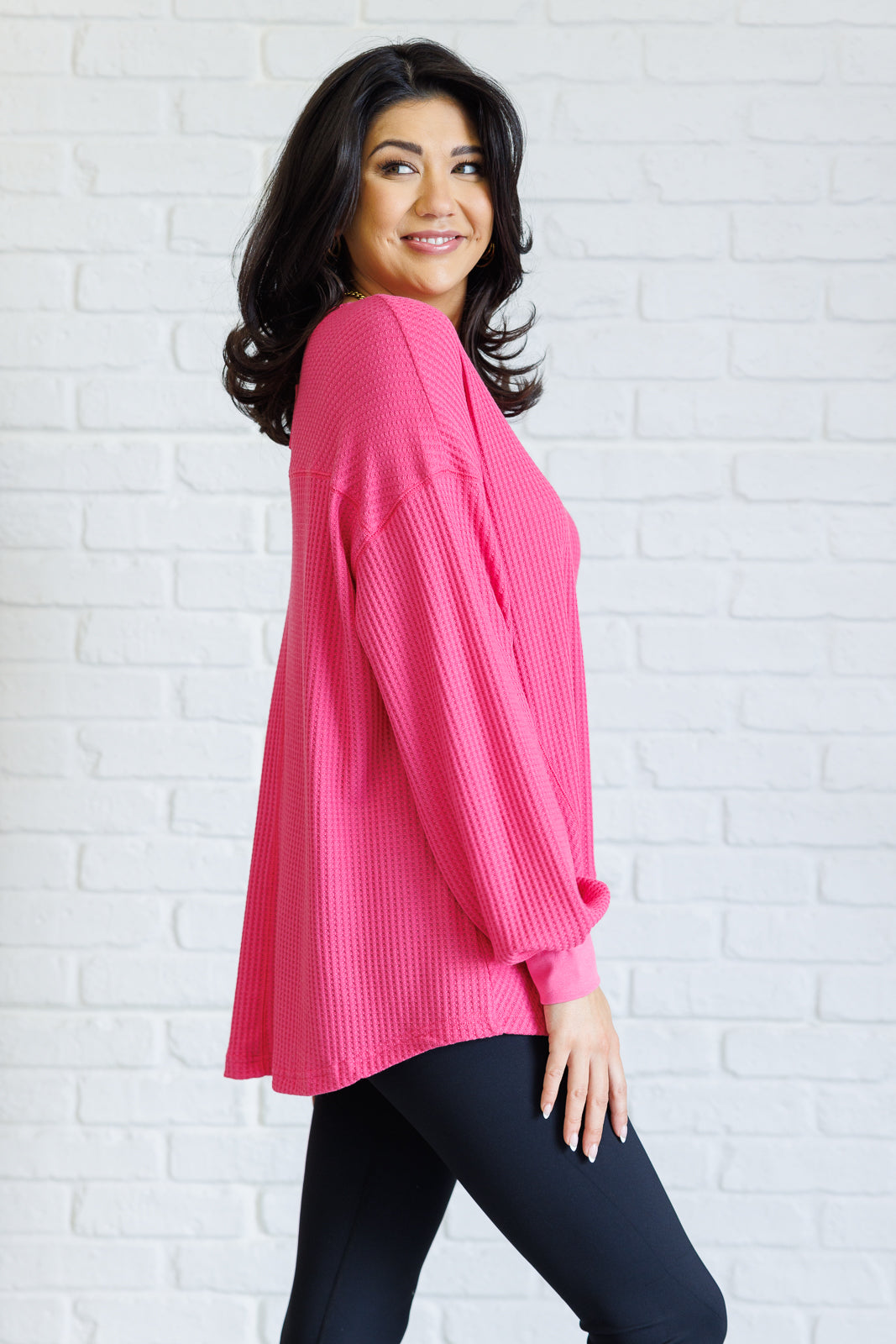 Women's pink waffle knit top with a V-neckline, long sleeves, banded sleeve cuffs, faux tag detail, and a scooped asymmetrical hemline.