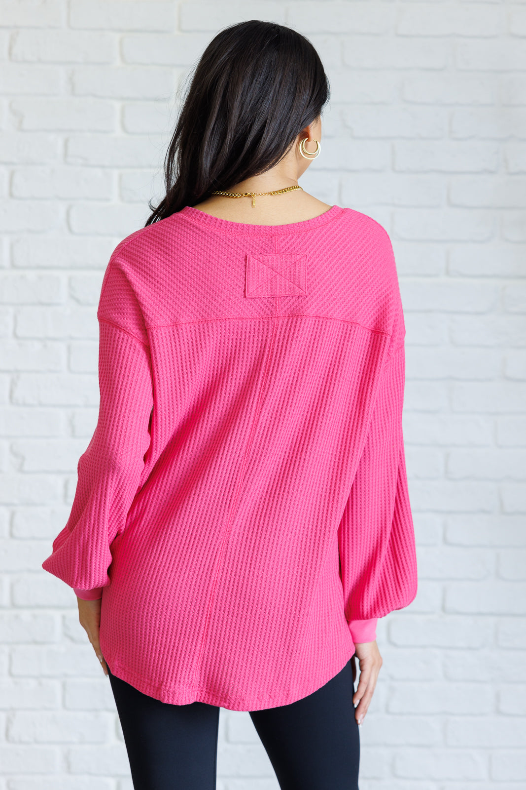 Women's pink waffle knit top with a V-neckline, long sleeves, banded sleeve cuffs, faux tag detail, and a scooped asymmetrical hemline.