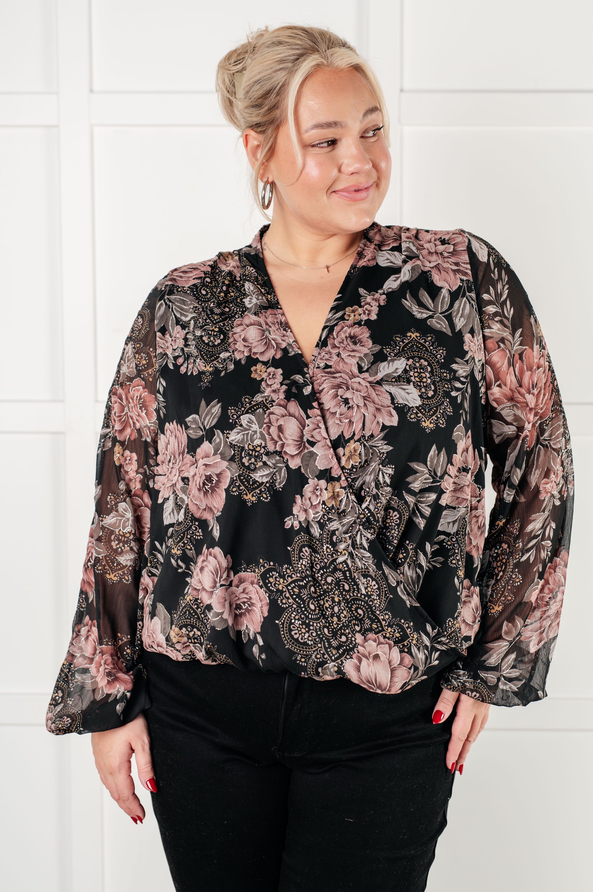 Plus size women's surplice floral blouse in black chiffon with floral print, featuring a v-neckline, surplice bodice with hook and eye closure, long balloon sleeves with cased elastic cuffs, and scooped hemline with cased elastic front hemline. Lined bodice.