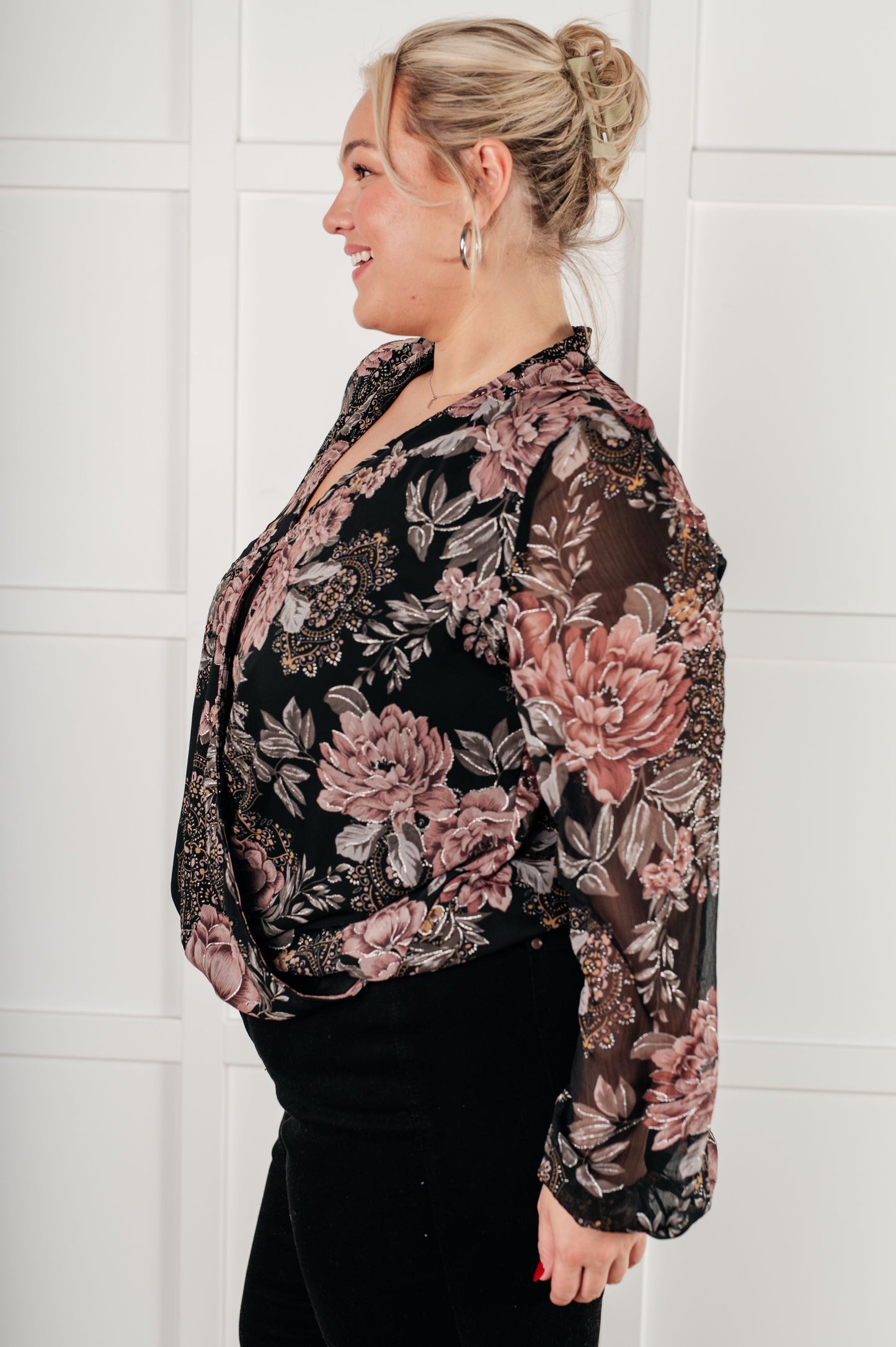 Side view of plus size surplice floral blouse in black chiffon with floral print, featuring a v-neckline, surplice bodice with hook and eye closure, long balloon sleeves with cased elastic cuffs, and scooped hemline with cased elastic front hemline. Lined bodice.