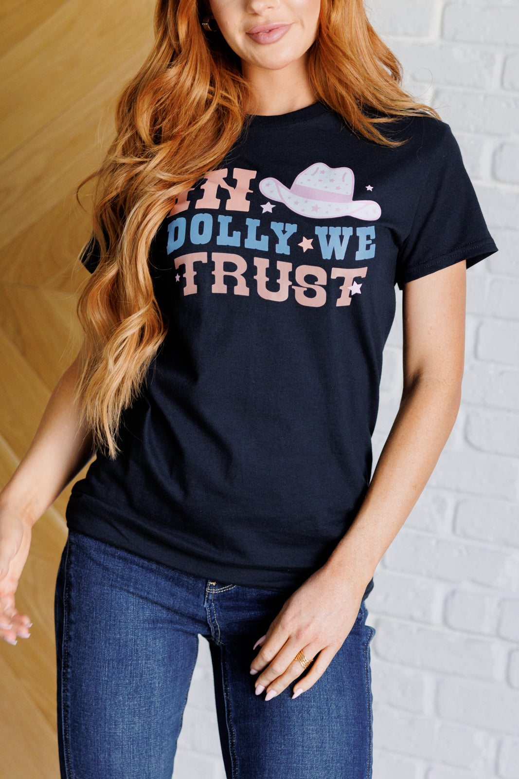 Graphic tee featuring 'In Dolly We Trust' slogan, made from soft jersey knit fabric. The design includes a ribbed crew neckline, short drop-shoulder sleeves, and a relaxed unisex fit. Made from 100% cotton.