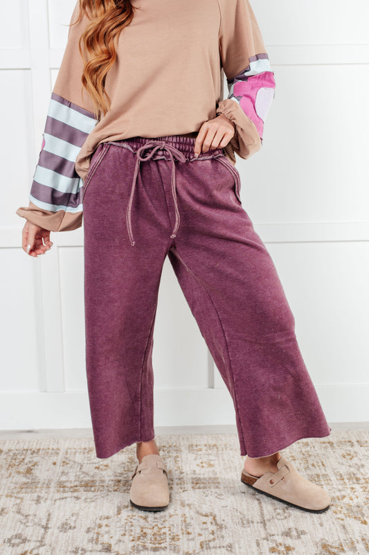 "Indie Wide Leg Cropped Pants in soft fleece, featuring a trendy mineral wash in eggplant. Designed with a drawstring elastic waist, functional side pockets, raw seam details, wide leg, and cropped length for a cozy, stylish, and relaxed look."