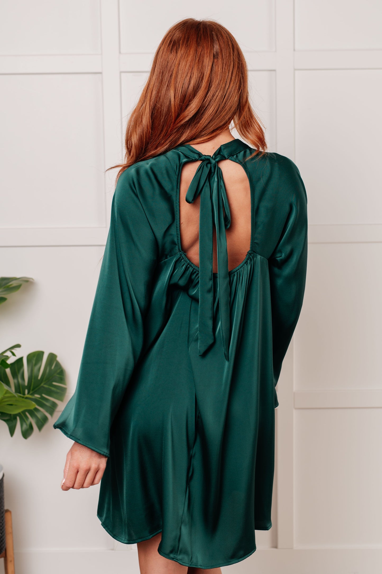 Emerald green poly satin A-line dress with banded mock neckline, long raglan bell sleeves, and back cased elastic for a flattering fit. Features a back cutout and tie closure. 