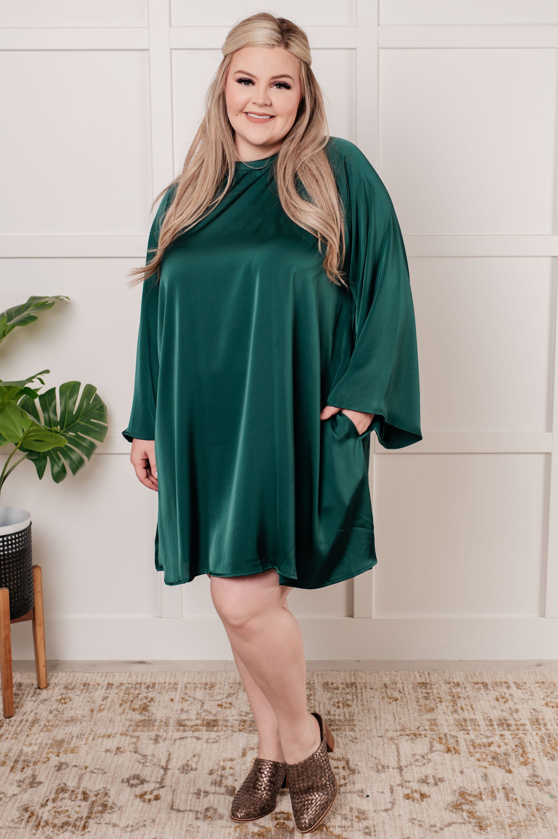 Emerald green poly satin A-line dress with banded mock neckline, long raglan bell sleeves, and back cased elastic for a flattering fit. Features a back cutout and tie closure. 