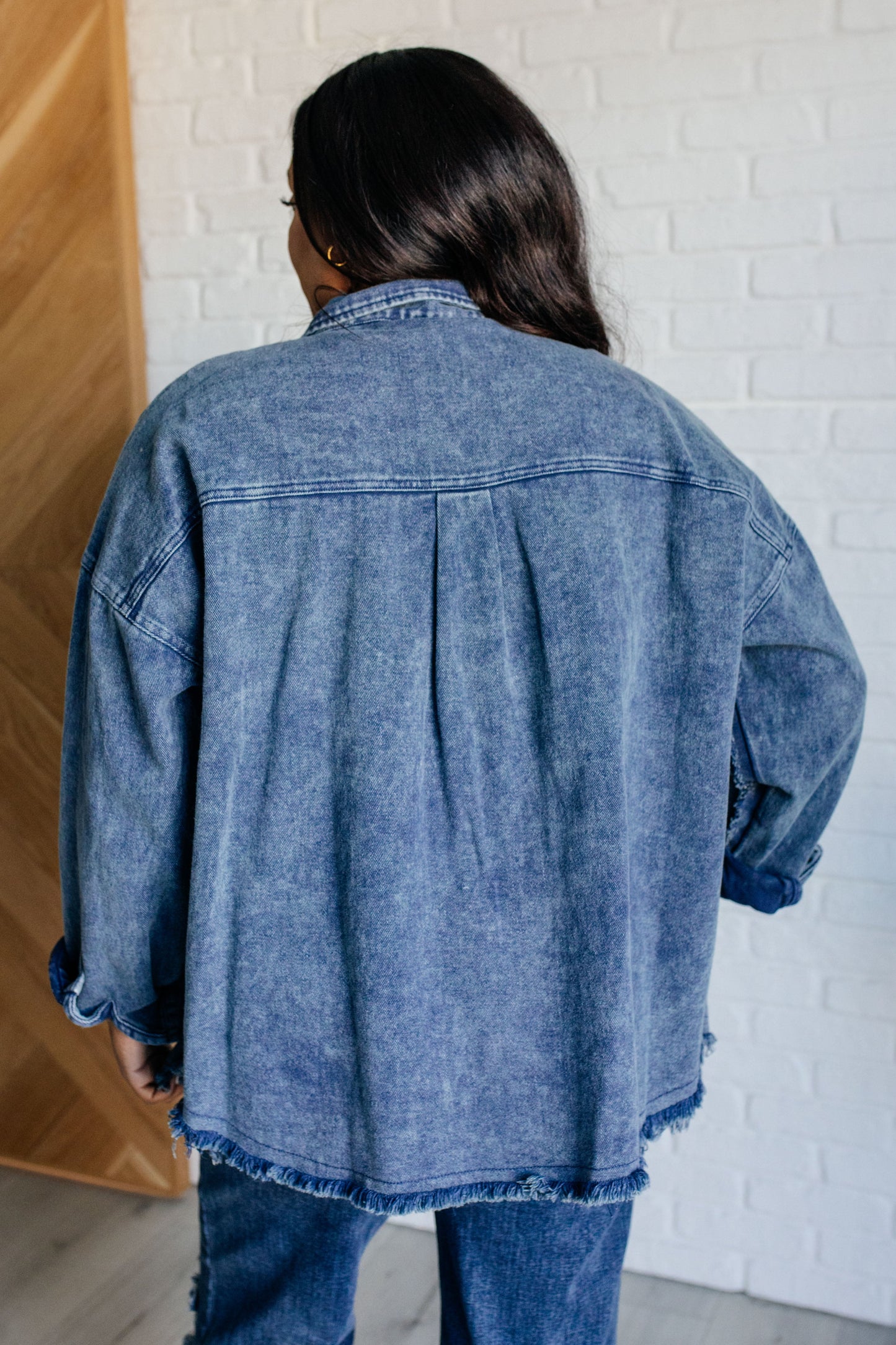 Dark wash mineral-wash denim shacket with a collared neckline, dropped shoulders, and long sleeves. Features a button front, gusseted pocket, raw edge details, back pleat, and raw hem with stay stitching.