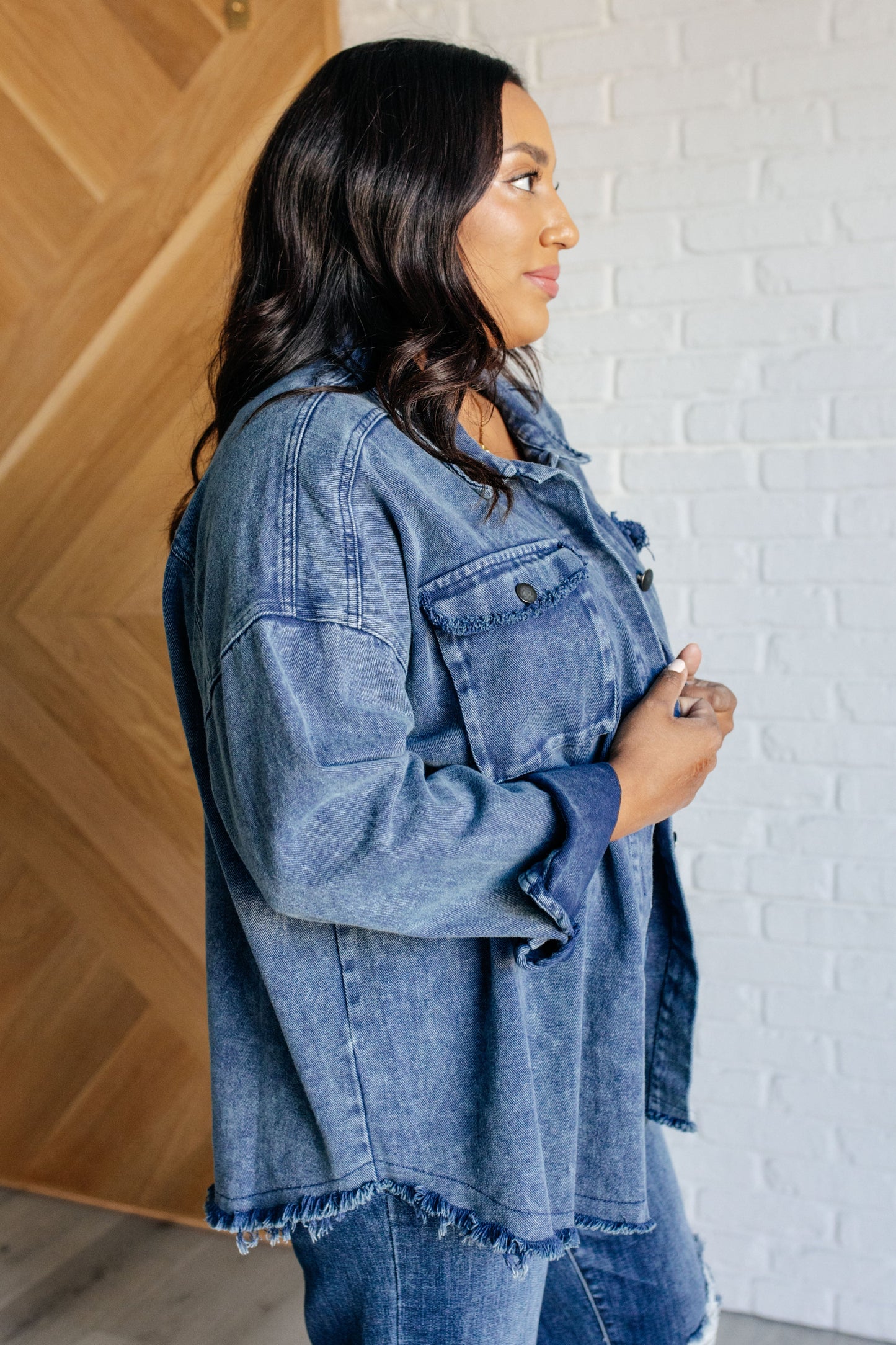 Dark wash mineral-wash denim shacket with a collared neckline, dropped shoulders, and long sleeves. Features a button front, gusseted pocket, raw edge details, back pleat, and raw hem with stay stitching.