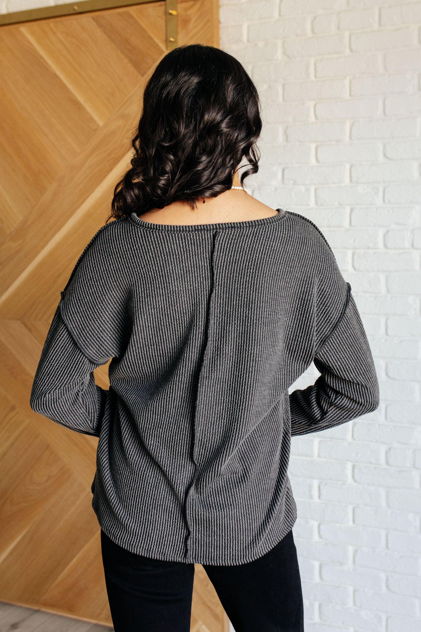 A women's ribbed knit top in charcoal, featuring a notch neckline, long drop-shoulder sleeves, a functional patch pocket, exposed seam details, and front slits.