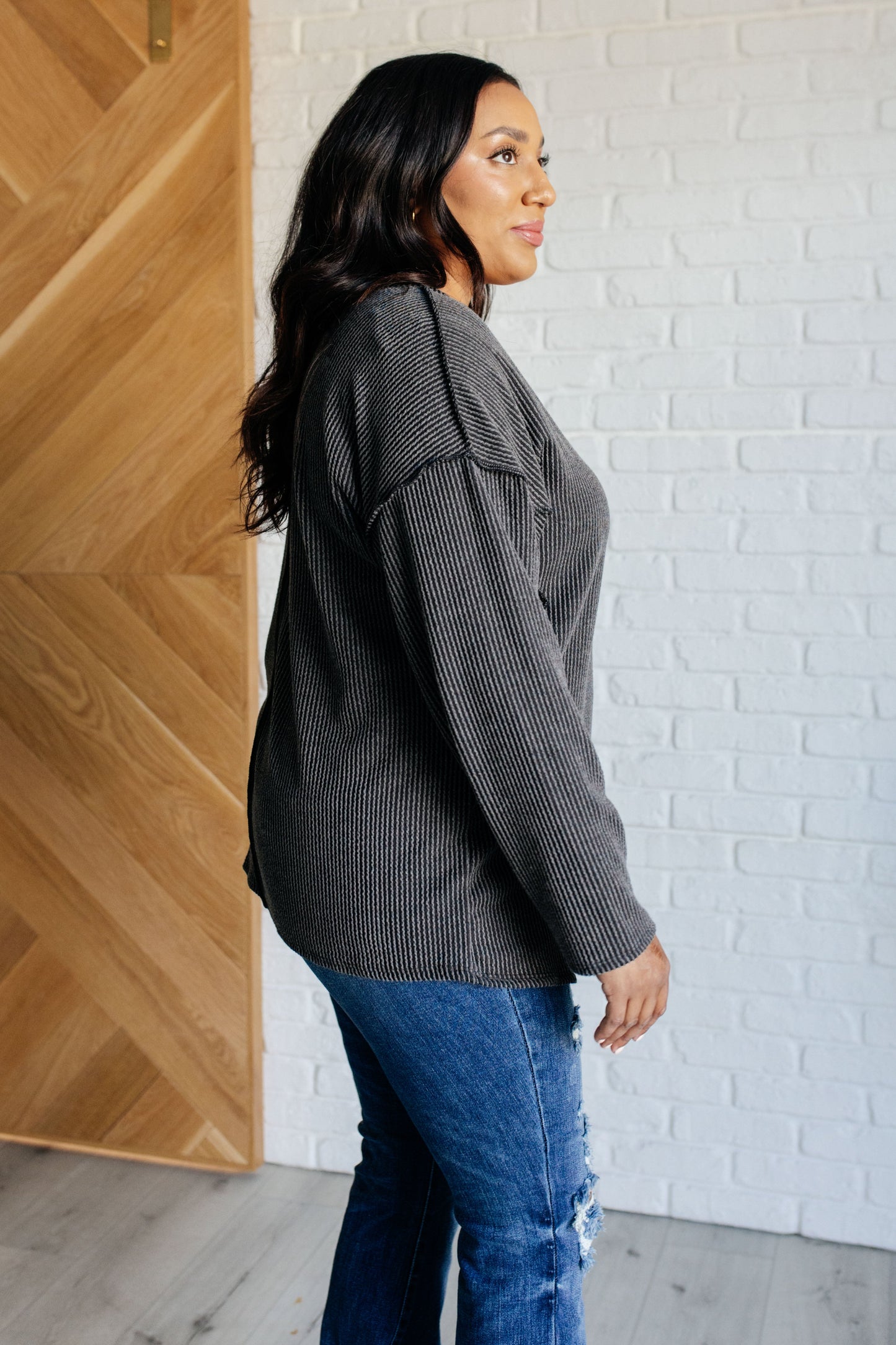 A women's ribbed knit top in charcoal, featuring a notch neckline, long drop-shoulder sleeves, a functional patch pocket, exposed seam details, and front slits.