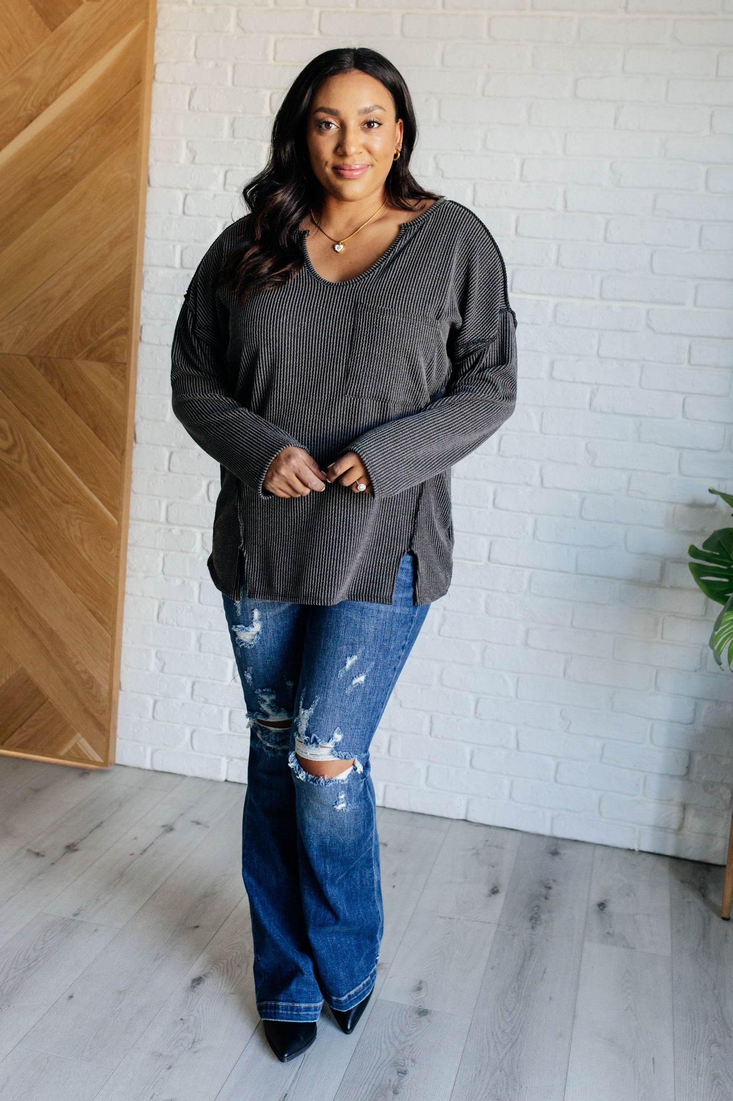 A women's ribbed knit top in charcoal, featuring a notch neckline, long drop-shoulder sleeves, a functional patch pocket, exposed seam details, and front slits.