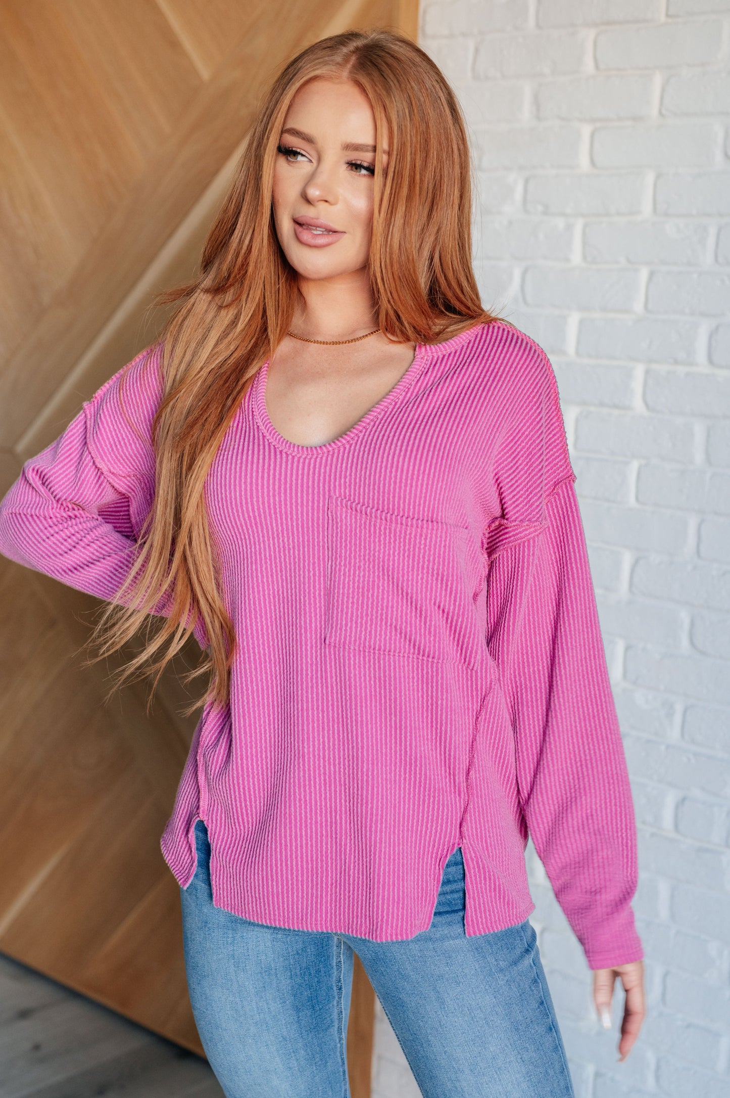 A women's ribbed knit top in magenta, featuring a notch neckline, long drop-shoulder sleeves, a functional patch pocket, exposed seam details, and front slits.