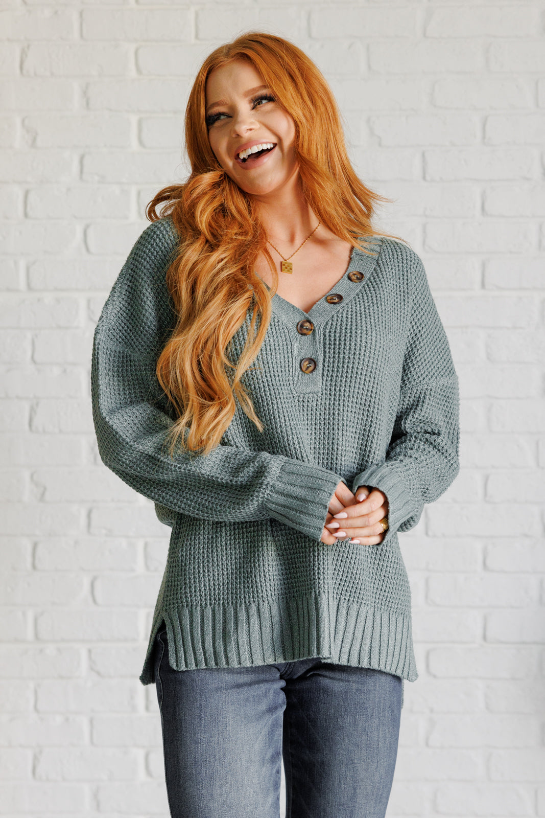 Women's sage waffle knit sweater with long sleeves, a banded ribbed V-neckline, exaggerated ribbed hemline and cuffs, side slits, and functional and faux button closures.