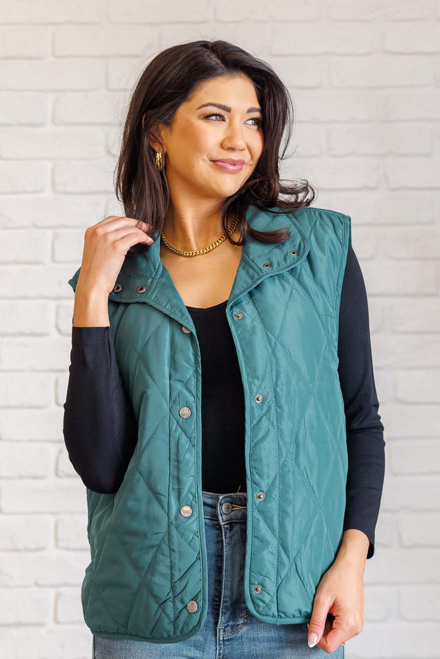 Hunter green puffer vest made from quilted windbreaker material. Features a high neckline, snap button closures, and functional side pockets. Fully lined with a round hemline.