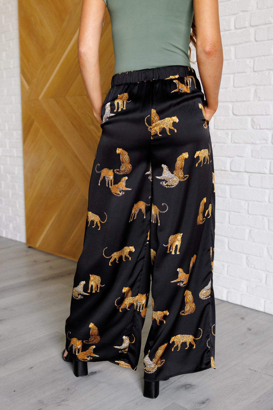 Satin Wide Leg Pants featuring a bold leopard print, high-rise elastic back waistband, and wide-leg silhouette. Made from poly satin with pleat front, pockets, and button-tab closure for a chic and comfortable fit.