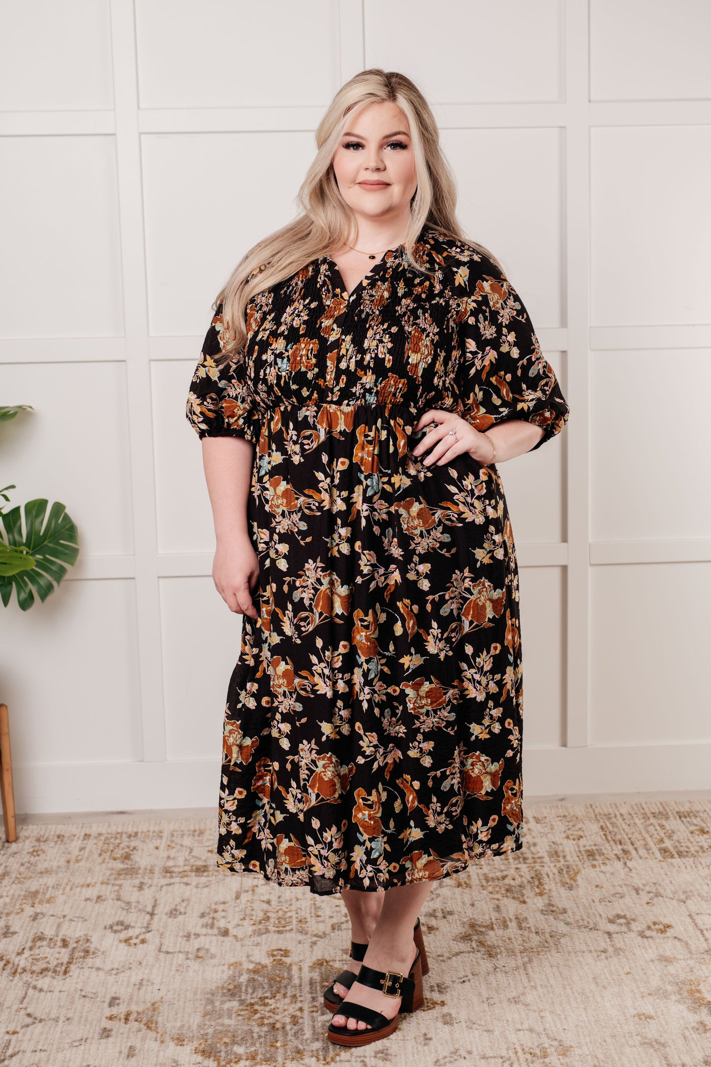 Black floral V-neck midi dress with smocked elastic bodice, button-down front, half balloon sleeves with cased elastic cuffs, and functional side pockets. Features ruffle detail and is fully lined.