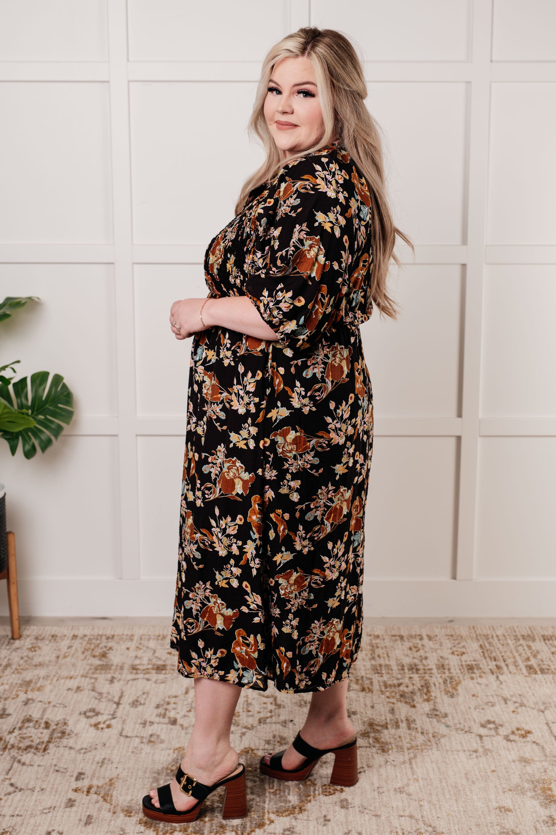 Black floral V-neck midi dress with smocked elastic bodice, button-down front, half balloon sleeves with cased elastic cuffs, and functional side pockets. Features ruffle detail and is fully lined.