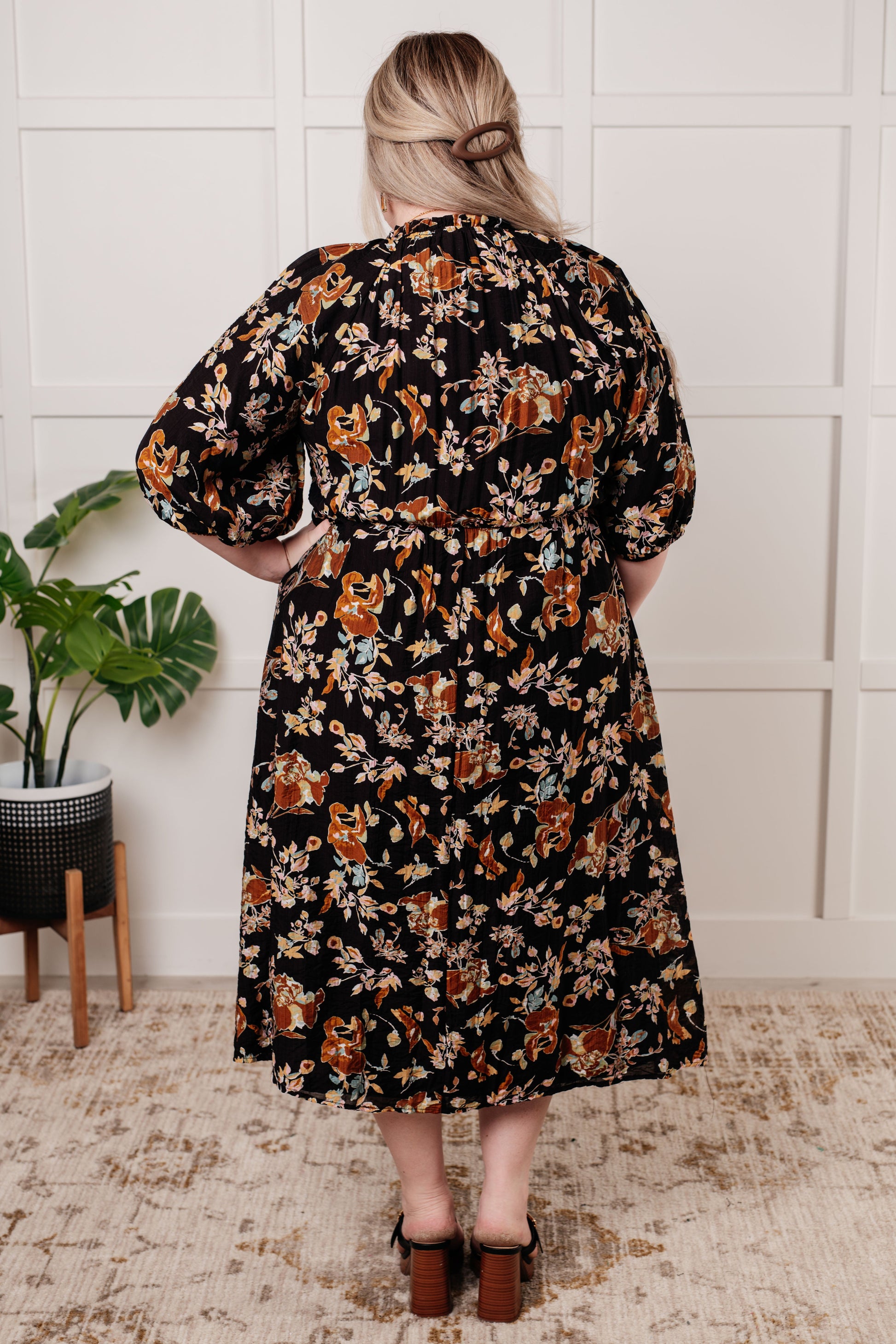 Black floral V-neck midi dress with smocked elastic bodice, button-down front, half balloon sleeves with cased elastic cuffs, and functional side pockets. Features ruffle detail and is fully lined.