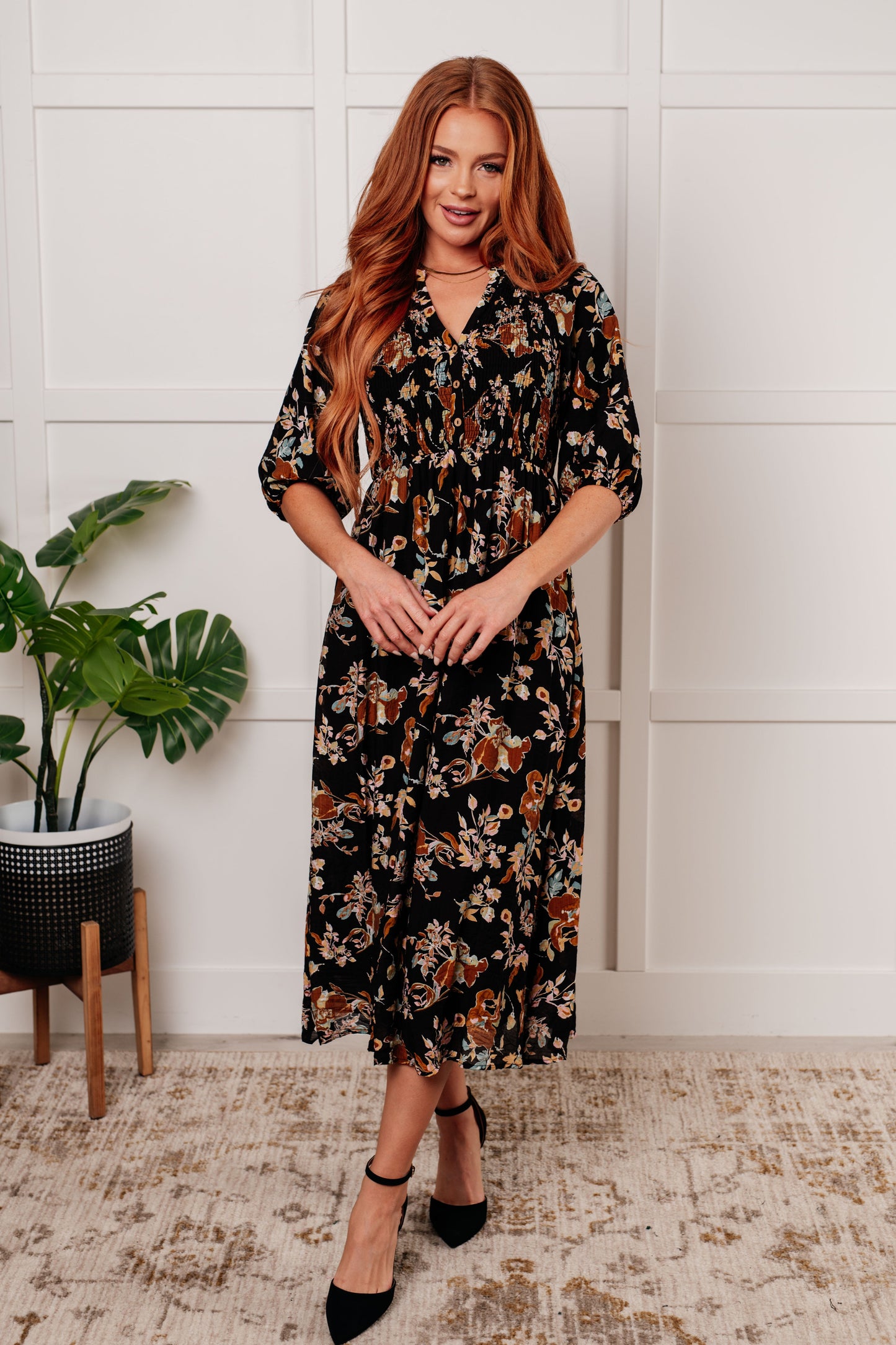 Black floral V-neck midi dress with smocked elastic bodice, button-down front, half balloon sleeves with cased elastic cuffs, and functional side pockets. Features ruffle detail and is fully lined.