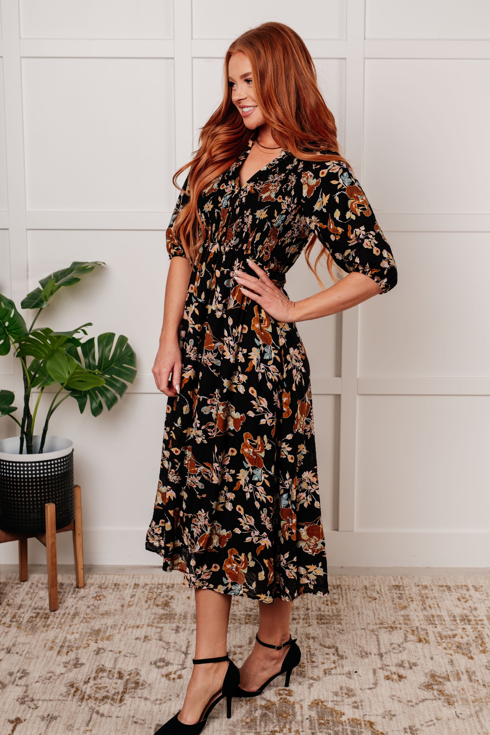 Black floral V-neck midi dress with smocked elastic bodice, button-down front, half balloon sleeves with cased elastic cuffs, and functional side pockets. Features ruffle detail and is fully lined.