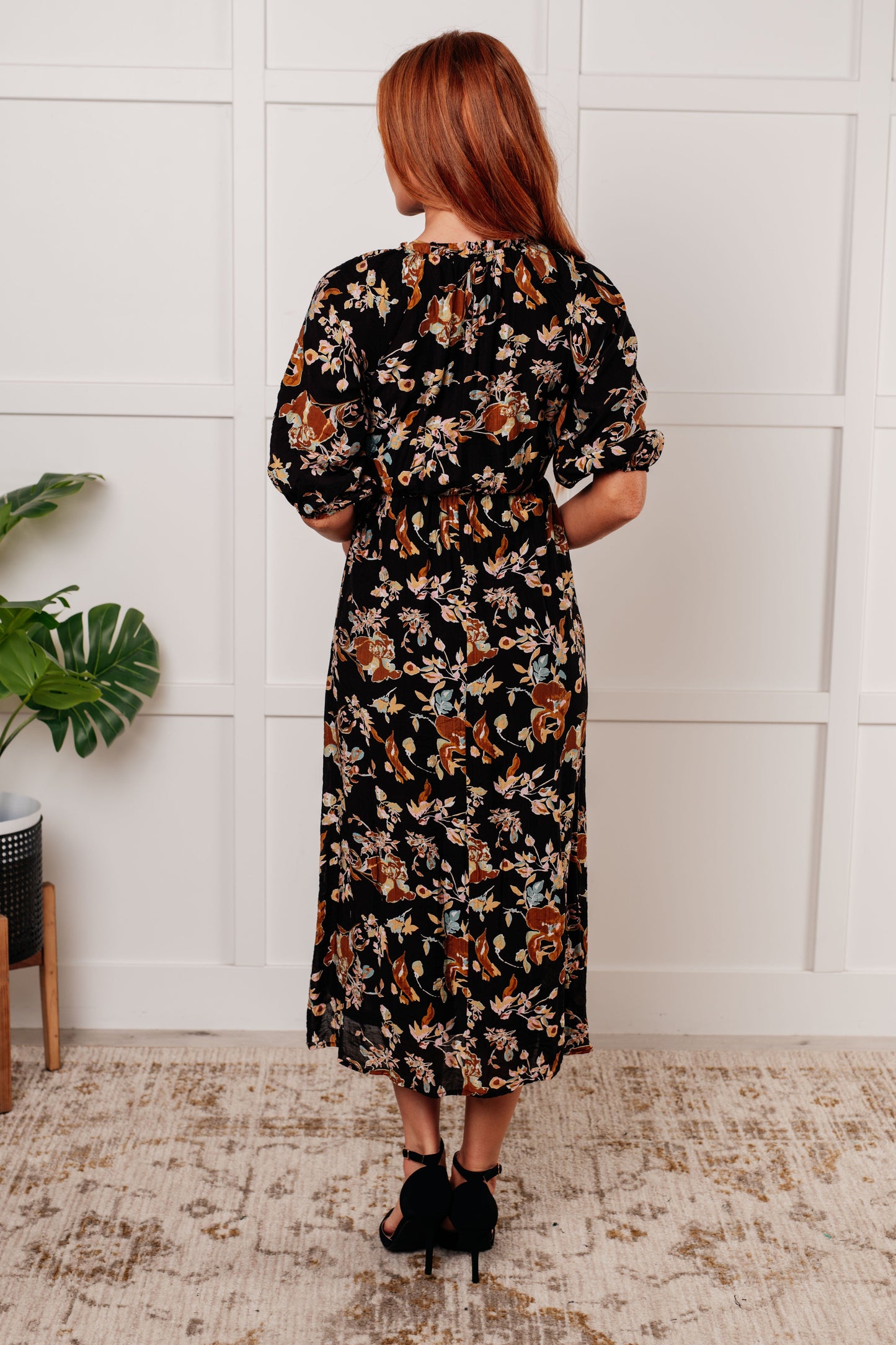 Black floral V-neck midi dress with smocked elastic bodice, button-down front, half balloon sleeves with cased elastic cuffs, and functional side pockets. Features ruffle detail and is fully lined.