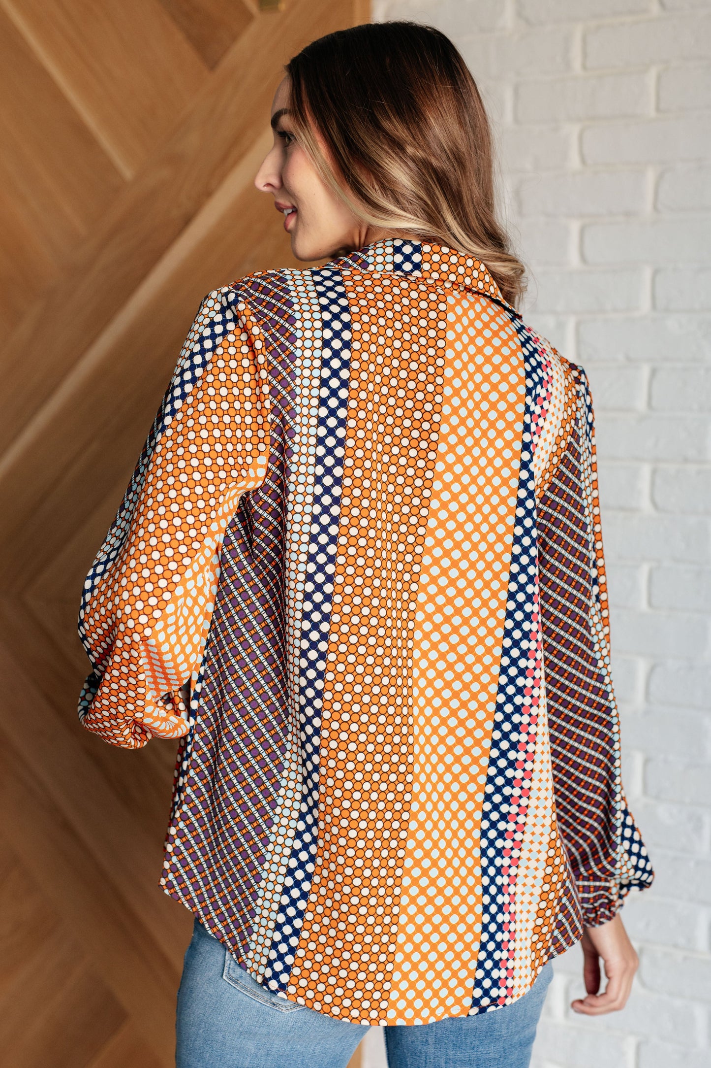 Navy, orange, brown, and pink polka dot poplin woven top with a collared neckline. Features long balloon sleeves with cased elastic cuffs, front button closures, and a scooped high-low hem.