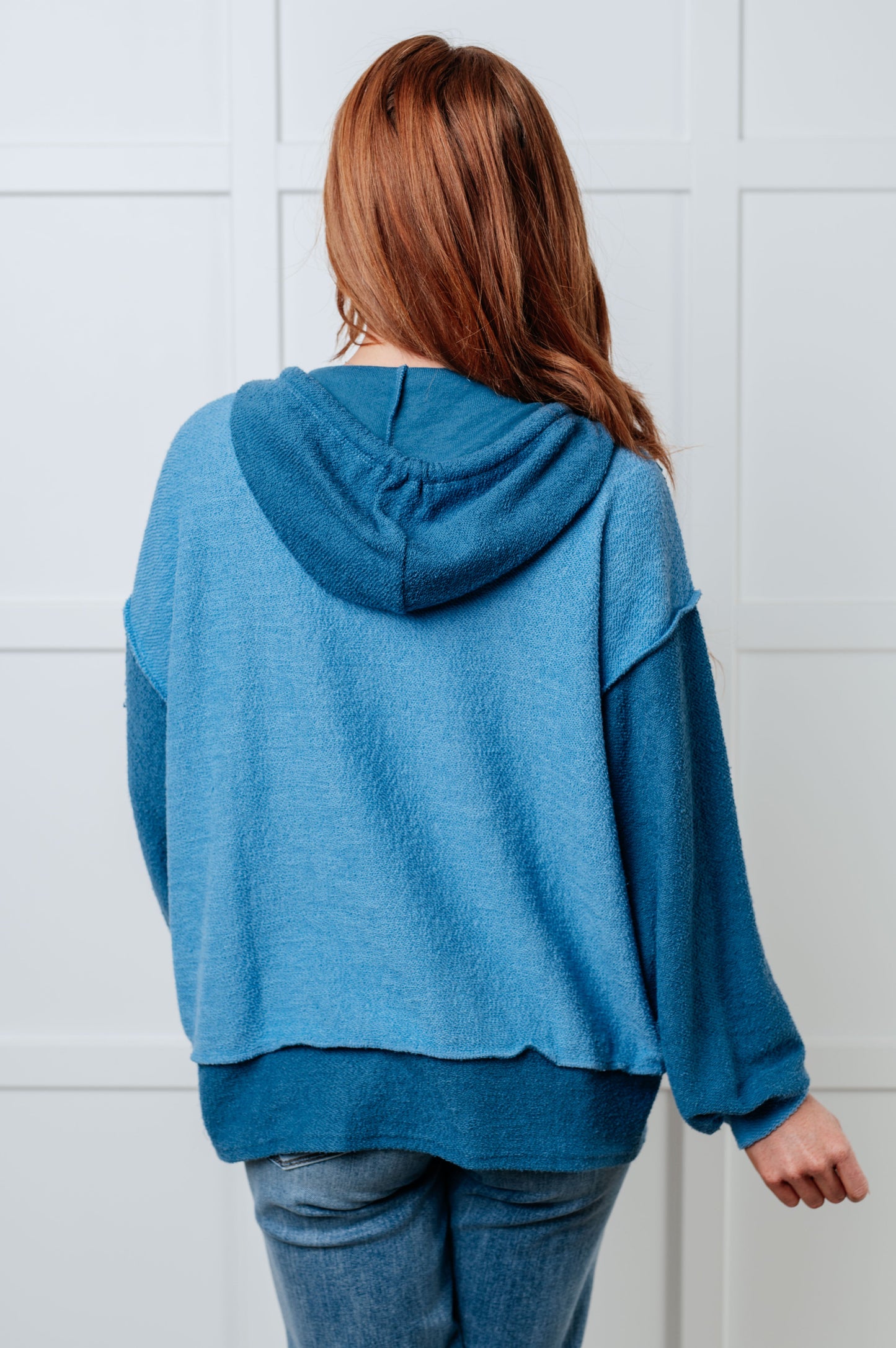 Women's blue hoodie with block color design, French Terry fabric, and hooded neckline with drawstring. Has long drop-shoulder sleeves, exposed seams, banded sleeve cuffs, and a stepped hem with side slits.