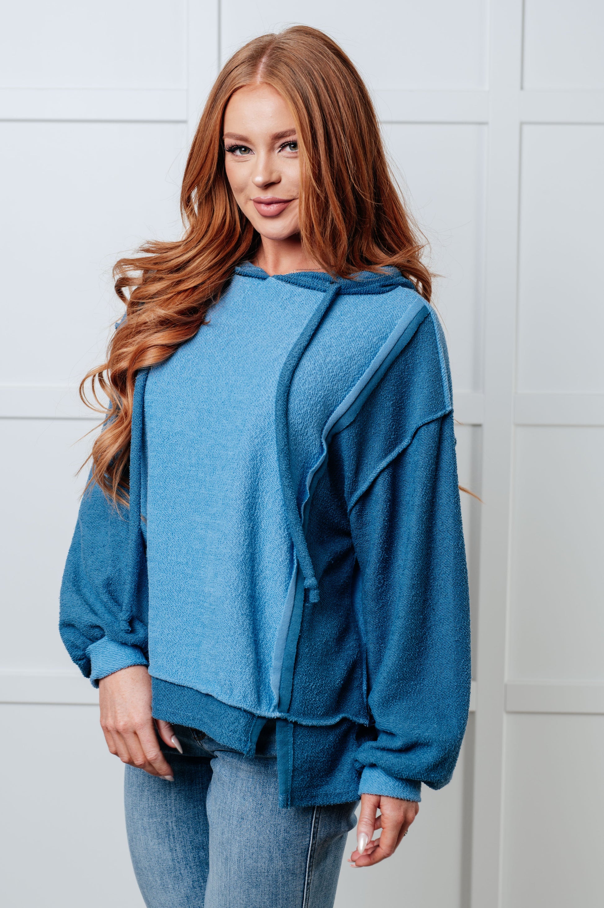 Women's blue hoodie with block color design, French Terry fabric, and hooded neckline with drawstring. Has long drop-shoulder sleeves, exposed seams, banded sleeve cuffs, and a stepped hem with side slits.