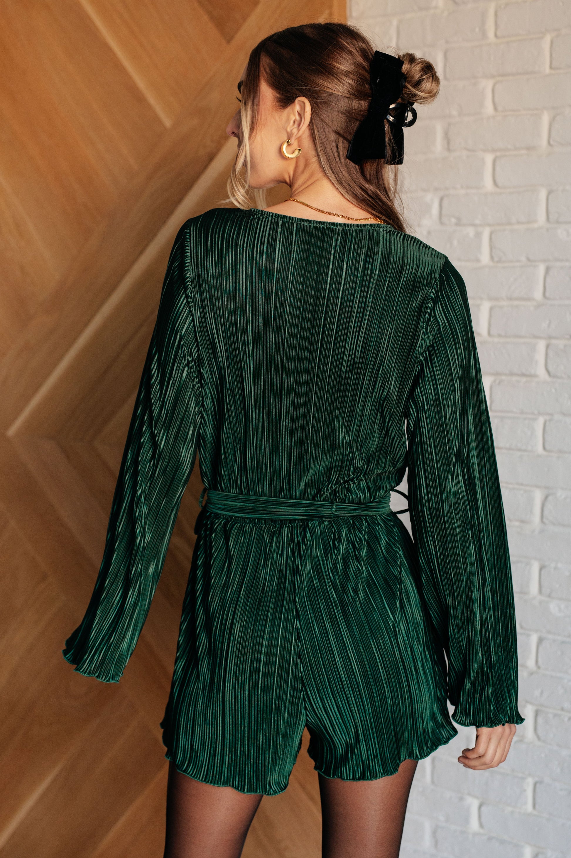 Dark green accordion plisse romper with a surplice front, V-neckline, and long sleeves. Features a self-tie belt, elastic waistband, and button closure.