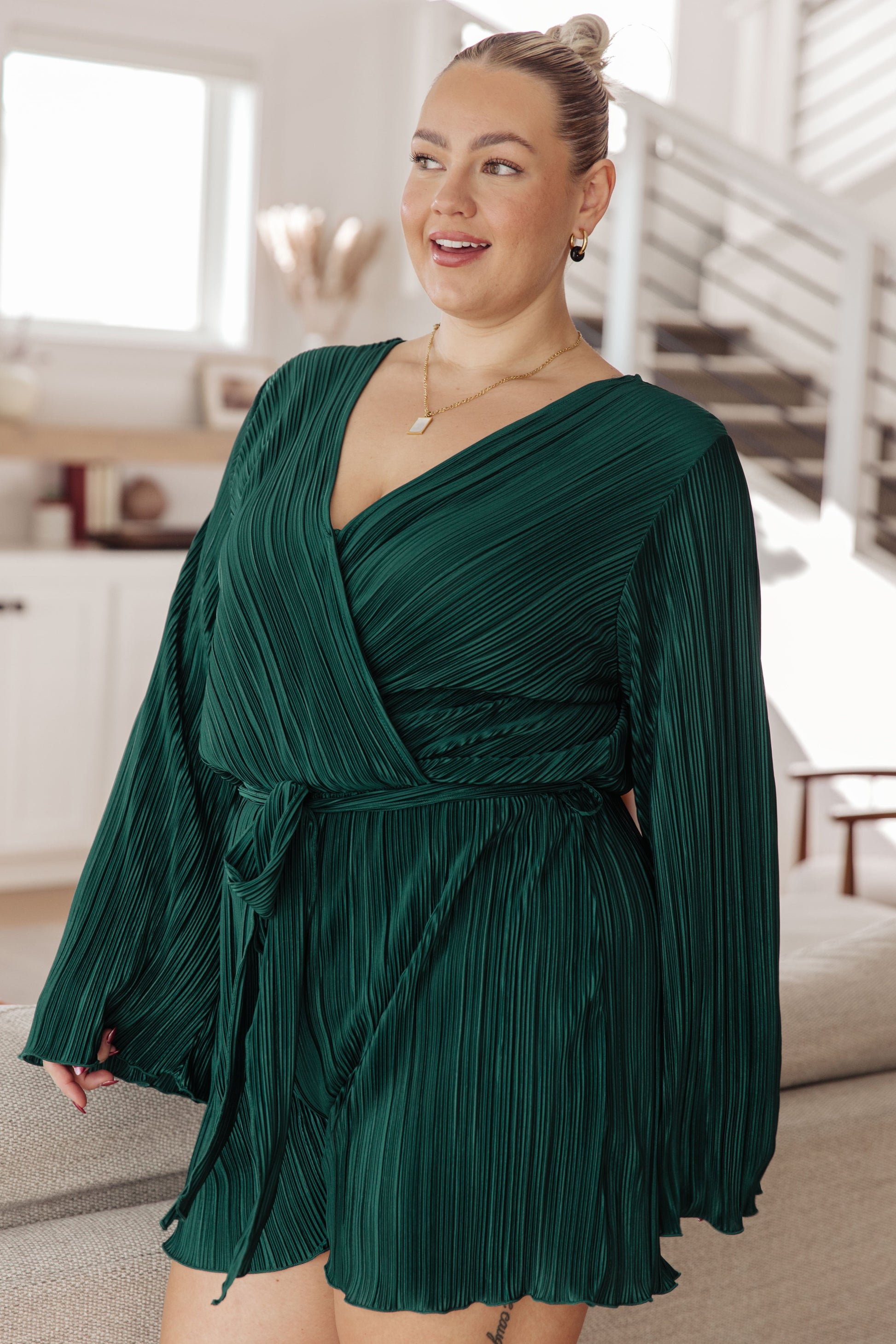 Dark green accordion plisse romper with a surplice front, V-neckline, and long sleeves. Features a self-tie belt, elastic waistband, and button closure.