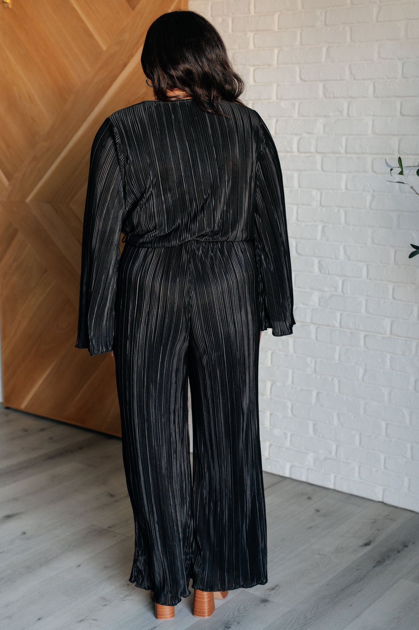 Black accordion plisse jumpsuit with a surplice front, V-neckline, and long sleeves. Features a self-tie belt, elastic waistband, and button closure.