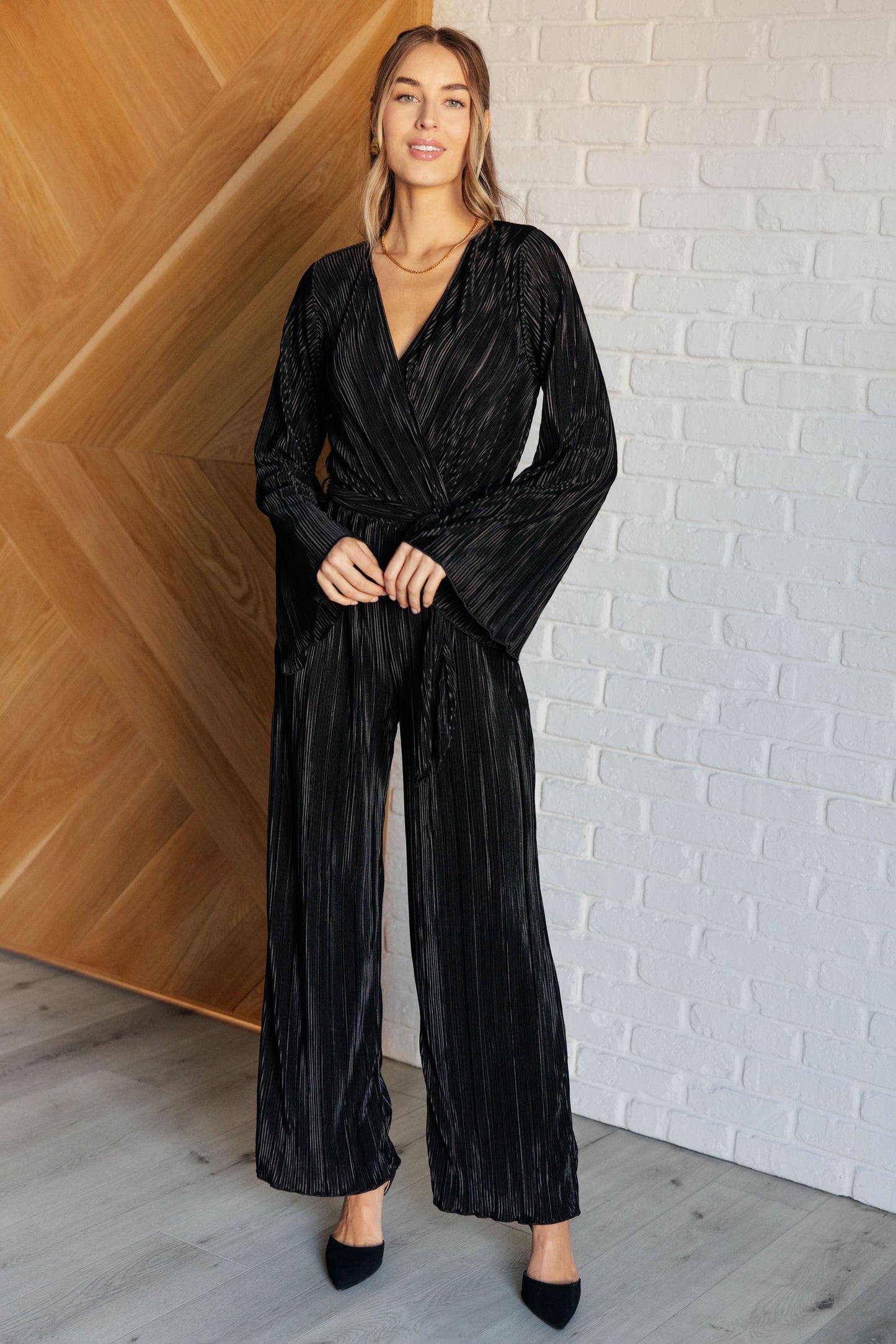 Black accordion plisse jumpsuit with a surplice front, V-neckline, and long sleeves. Features a self-tie belt, elastic waistband, and button closure.