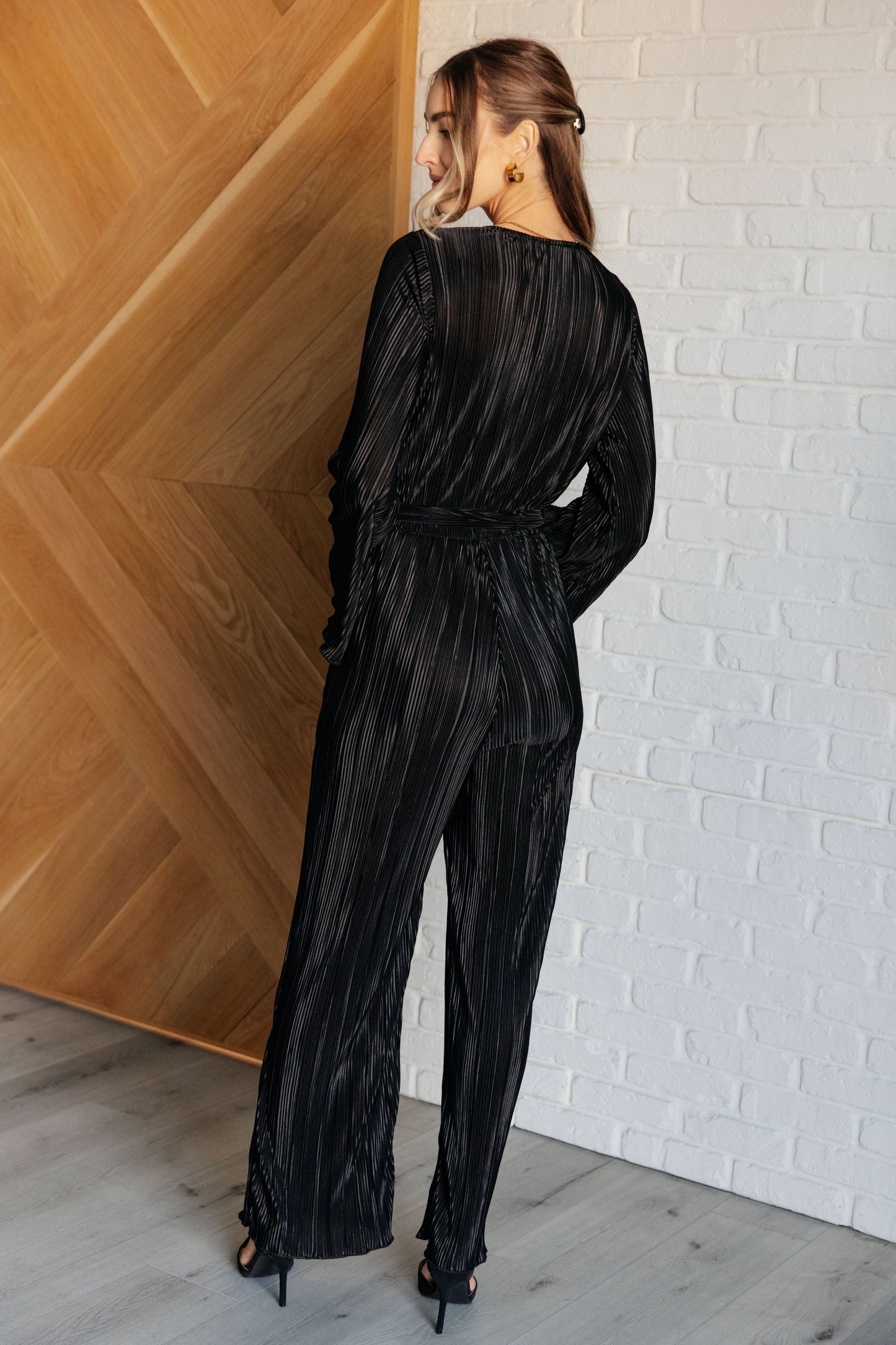 Black accordion plisse jumpsuit with a surplice front, V-neckline, and long sleeves. Features a self-tie belt, elastic waistband, and button closure.