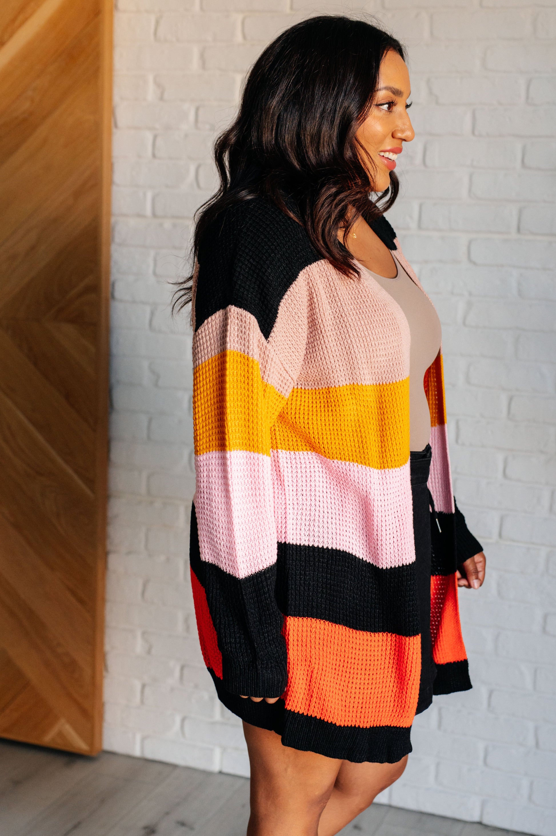 Striped open-front sweater with a cozy knit fabric featuring beige, pink, yellow, black, and orange stripes. Designed with long drop-shoulder sleeves and banded ribbed cuffs and hemline for a relaxed fit.