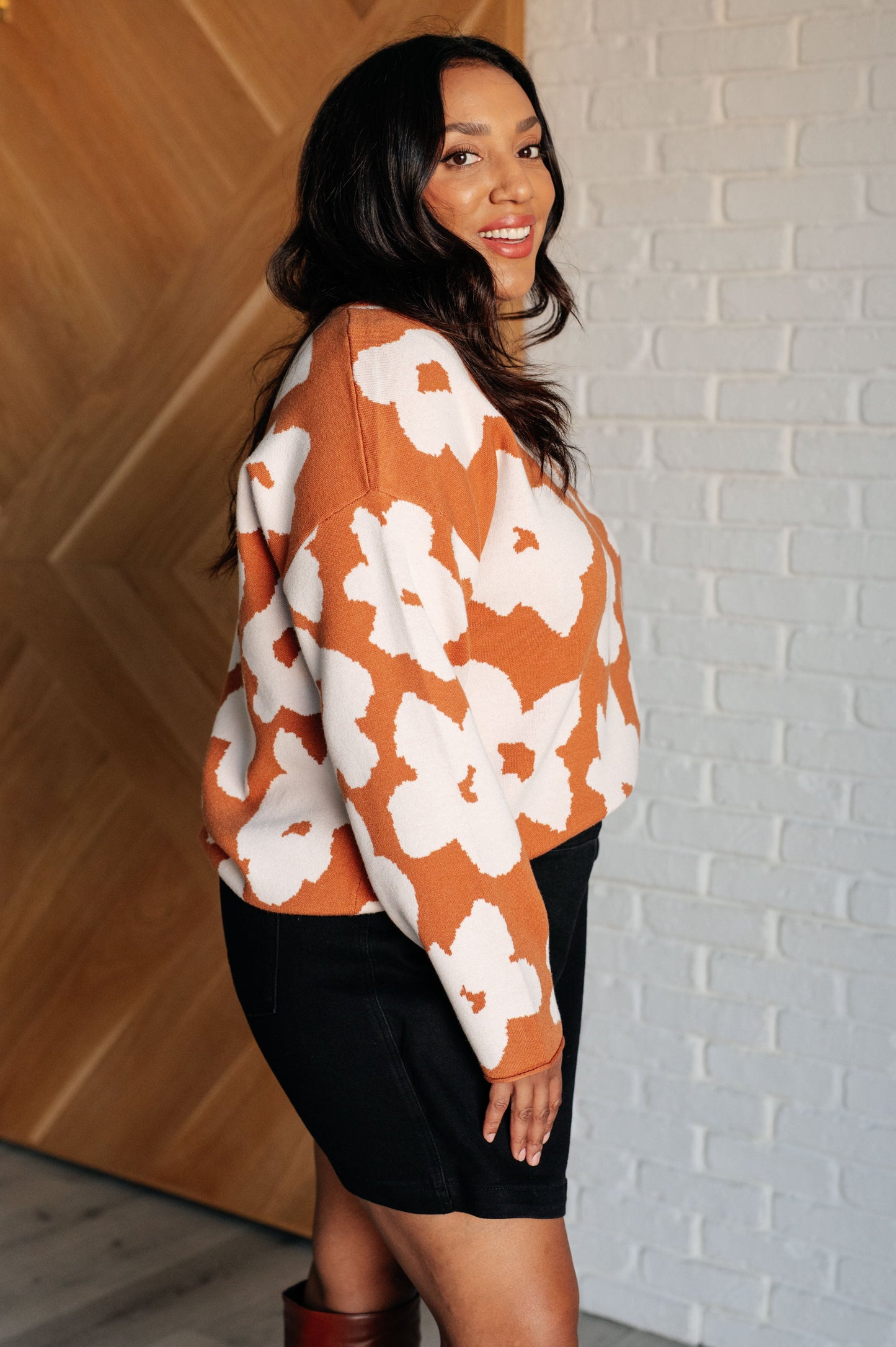 A rust-colored sweater made from mid-weight knit fabric featuring a white floral print. The design includes a round neckline, long drop-shoulder sleeves, and a relaxed fit silhouette.