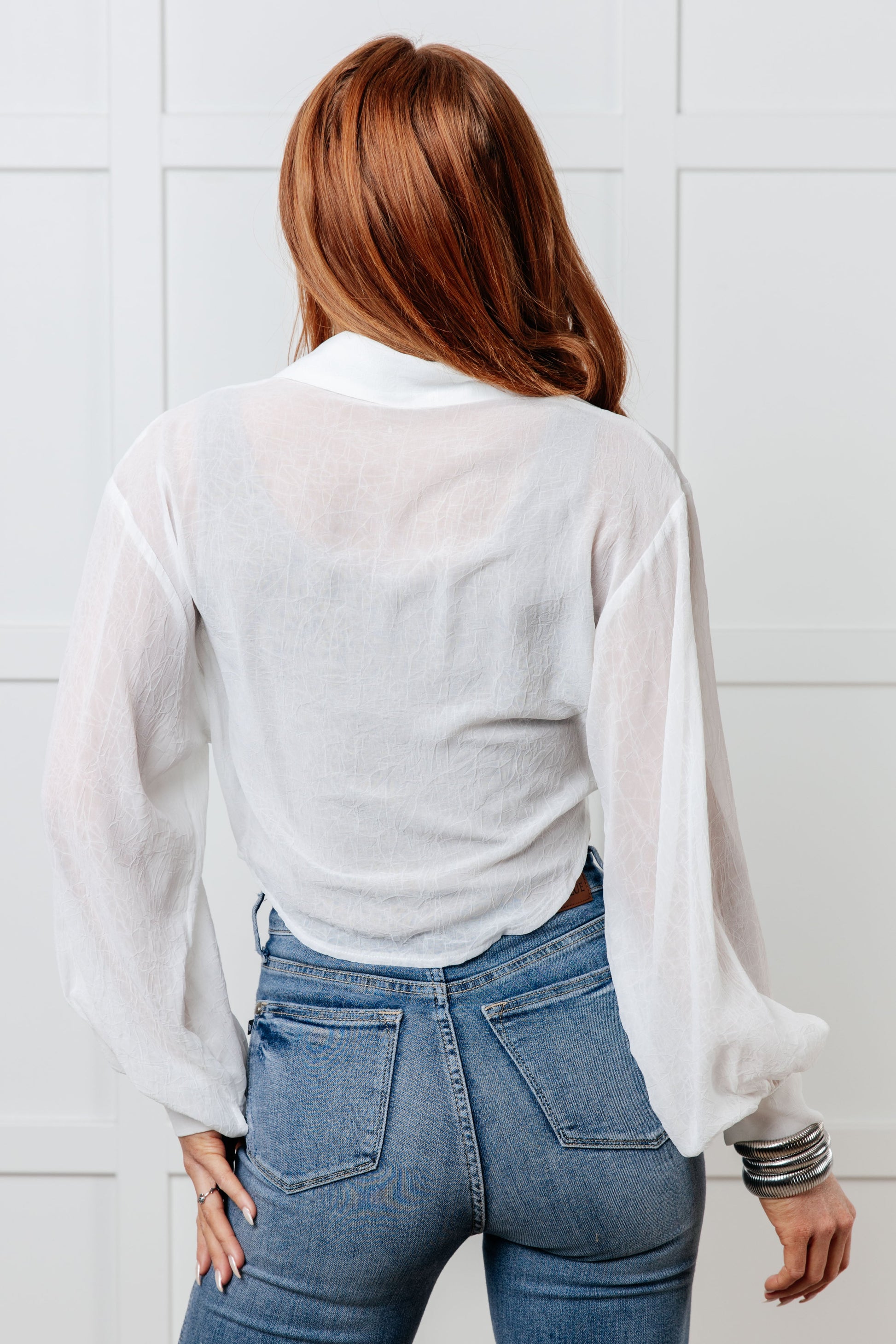 A women's button-up top made from crinkle woven fabric, featuring a collared neckline, long balloon sleeves with banded button cuffs, full button closures, a waist tie, a cropped length, and a scooped hem.