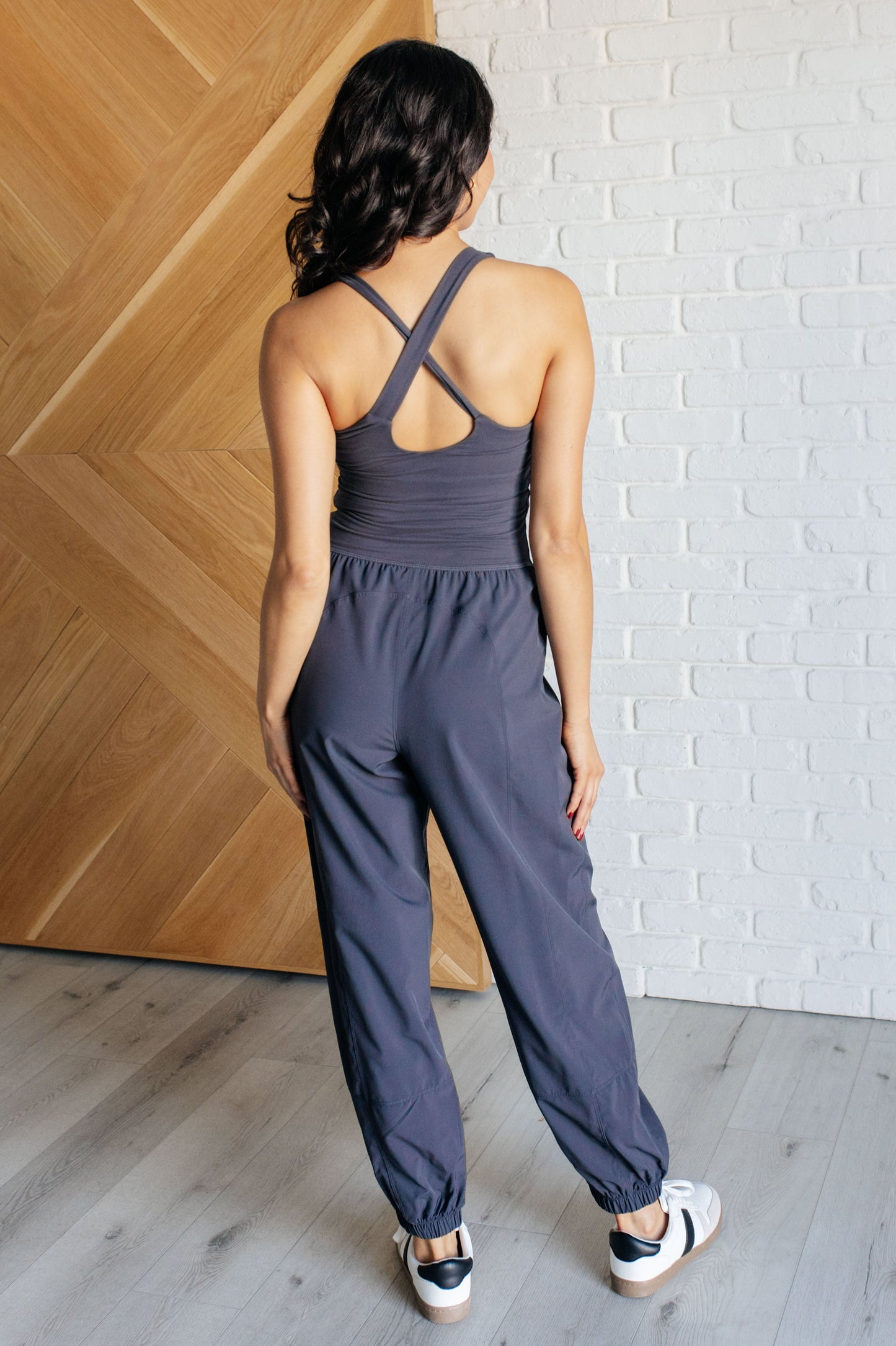 Charcoal jumpsuit with a square neckline, sleeveless design, crossed back straps, cutout detail, jogger-style silhouette, built-in shelf bra with removable cups, and functional side pockets.