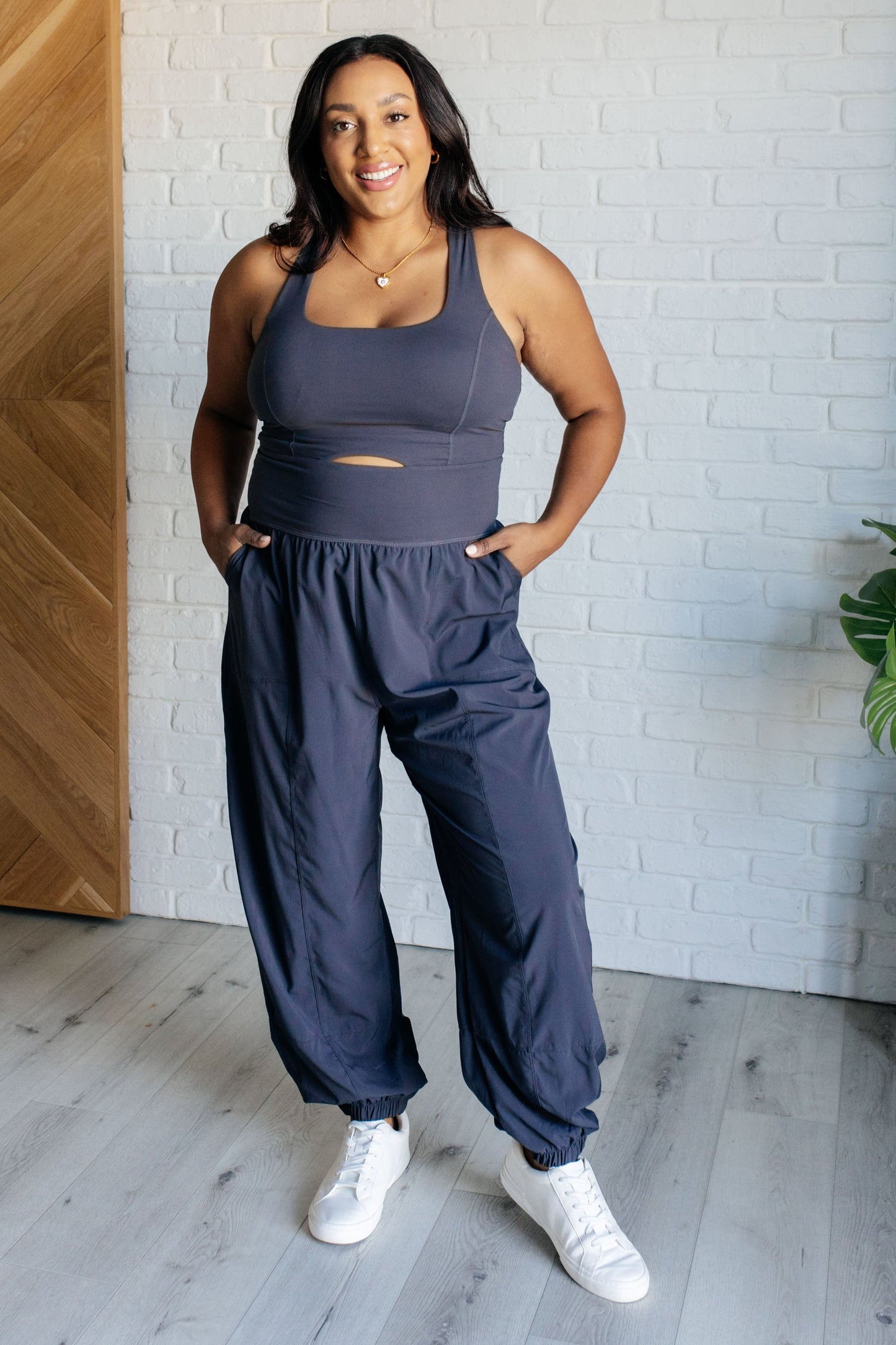 Charcoal jumpsuit with a square neckline, sleeveless design, crossed back straps, cutout detail, jogger-style silhouette, built-in shelf bra with removable cups, and functional side pockets.