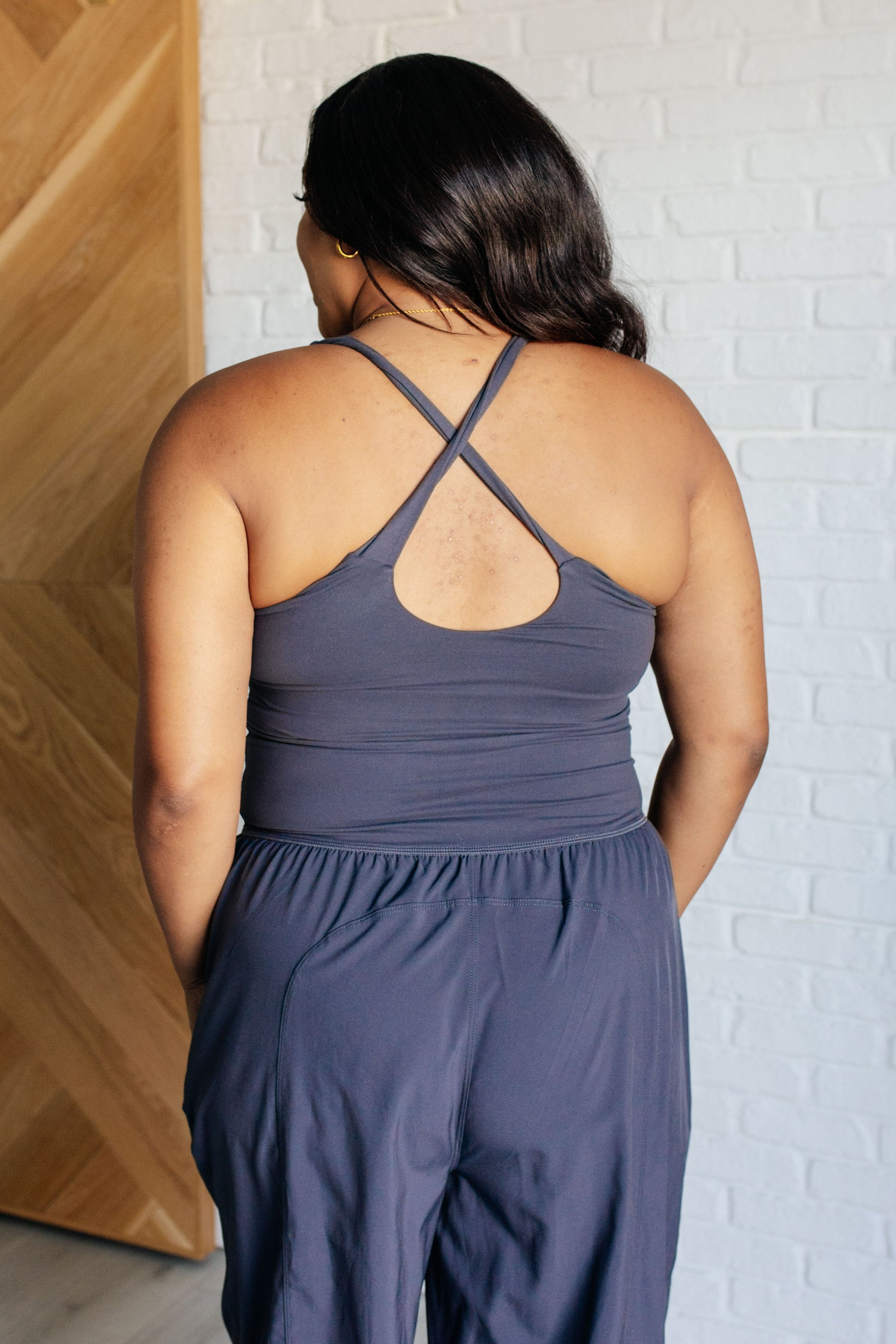 Charcoal jumpsuit with a square neckline, sleeveless design, crossed back straps, cutout detail, jogger-style silhouette, built-in shelf bra with removable cups, and functional side pockets.