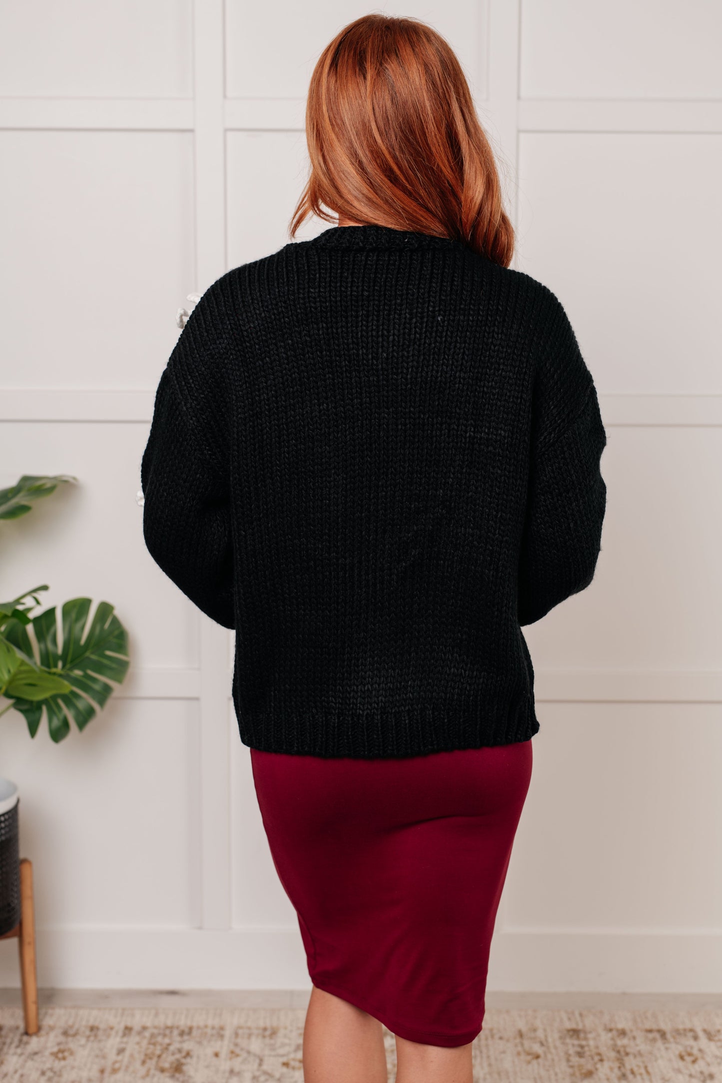 A black open-front sweater knit cardigan featuring long balloon sleeves with drop shoulders. The design includes white yarn bow details, along with ribbed sleeve cuffs, hem, and neckline.