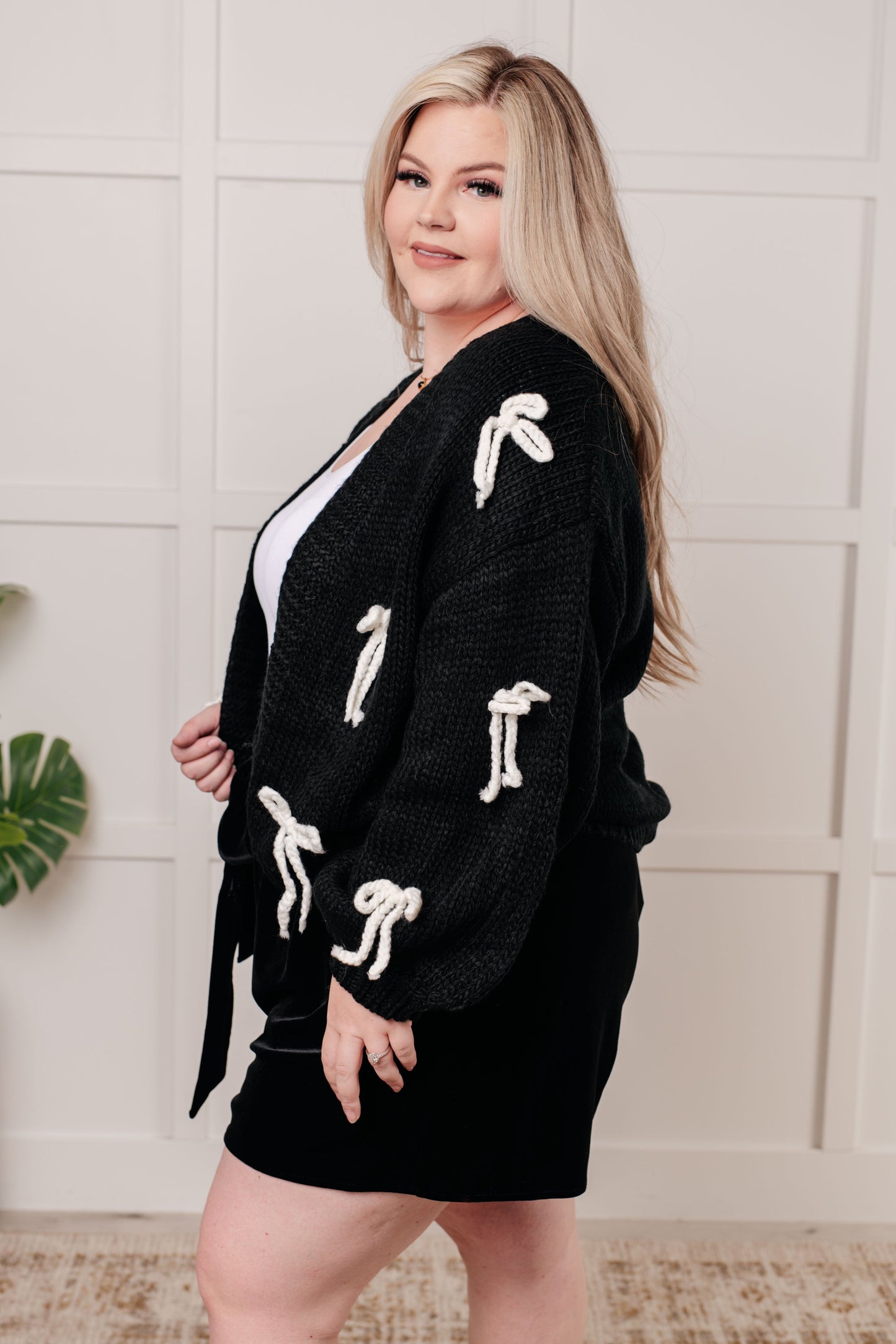 A black open-front sweater knit cardigan featuring long balloon sleeves with drop shoulders. The design includes white yarn bow details, along with ribbed sleeve cuffs, hem, and neckline.