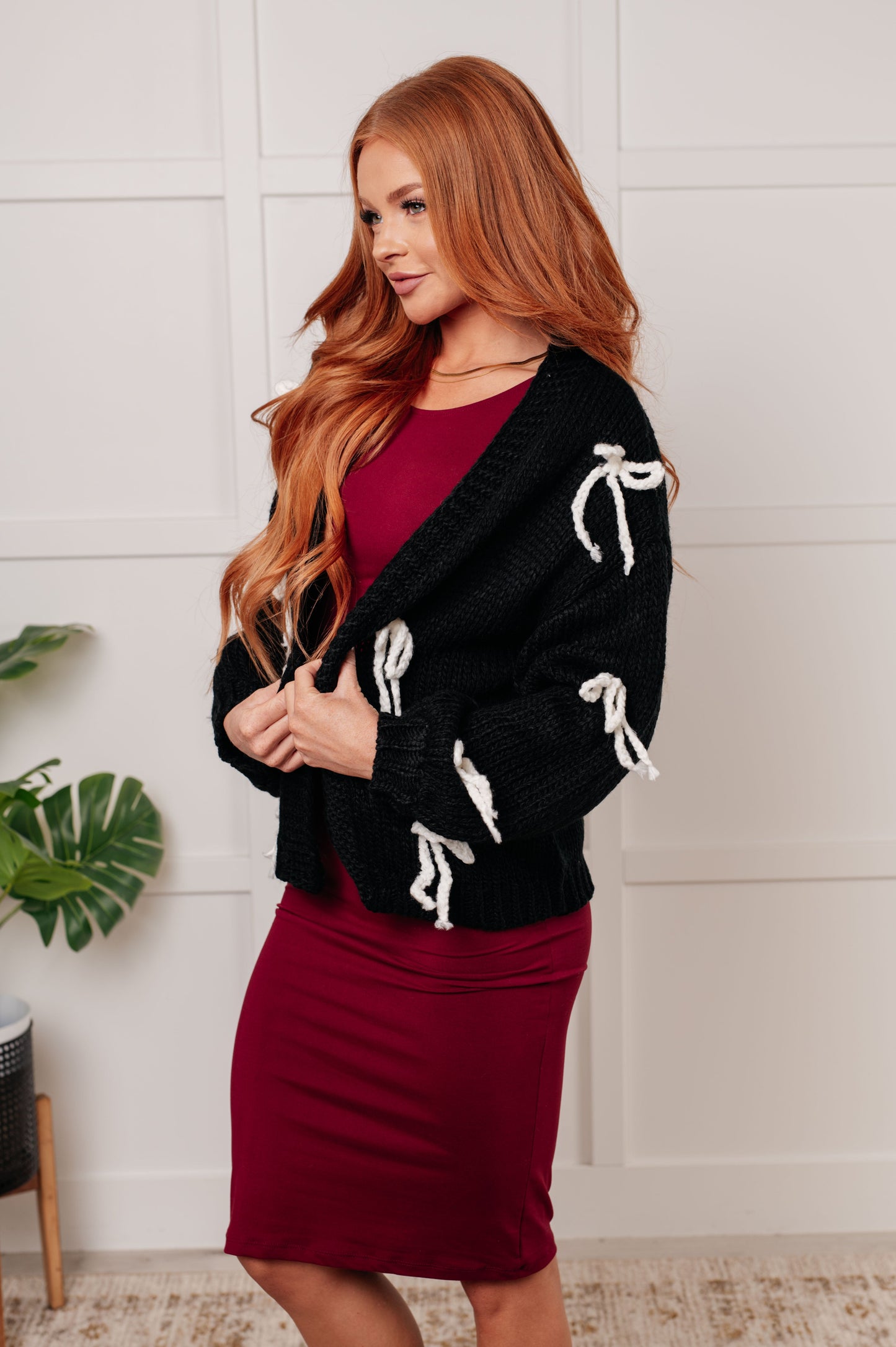 A black open-front sweater knit cardigan featuring long balloon sleeves with drop shoulders. The design includes white yarn bow details, along with ribbed sleeve cuffs, hem, and neckline.